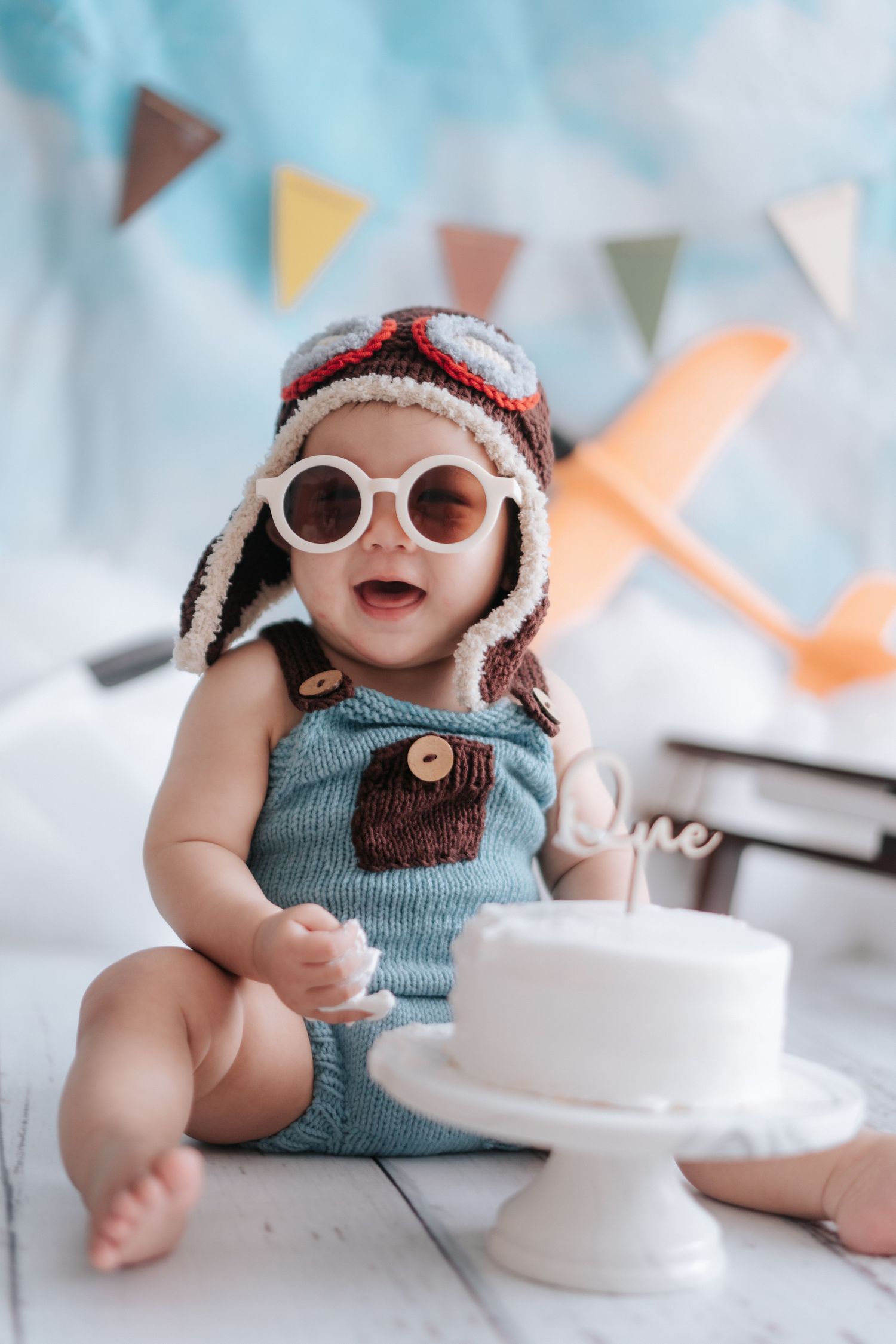 A small doll wearing a blue knit top and white sunglasses sits against a pastel background with festive bunting.