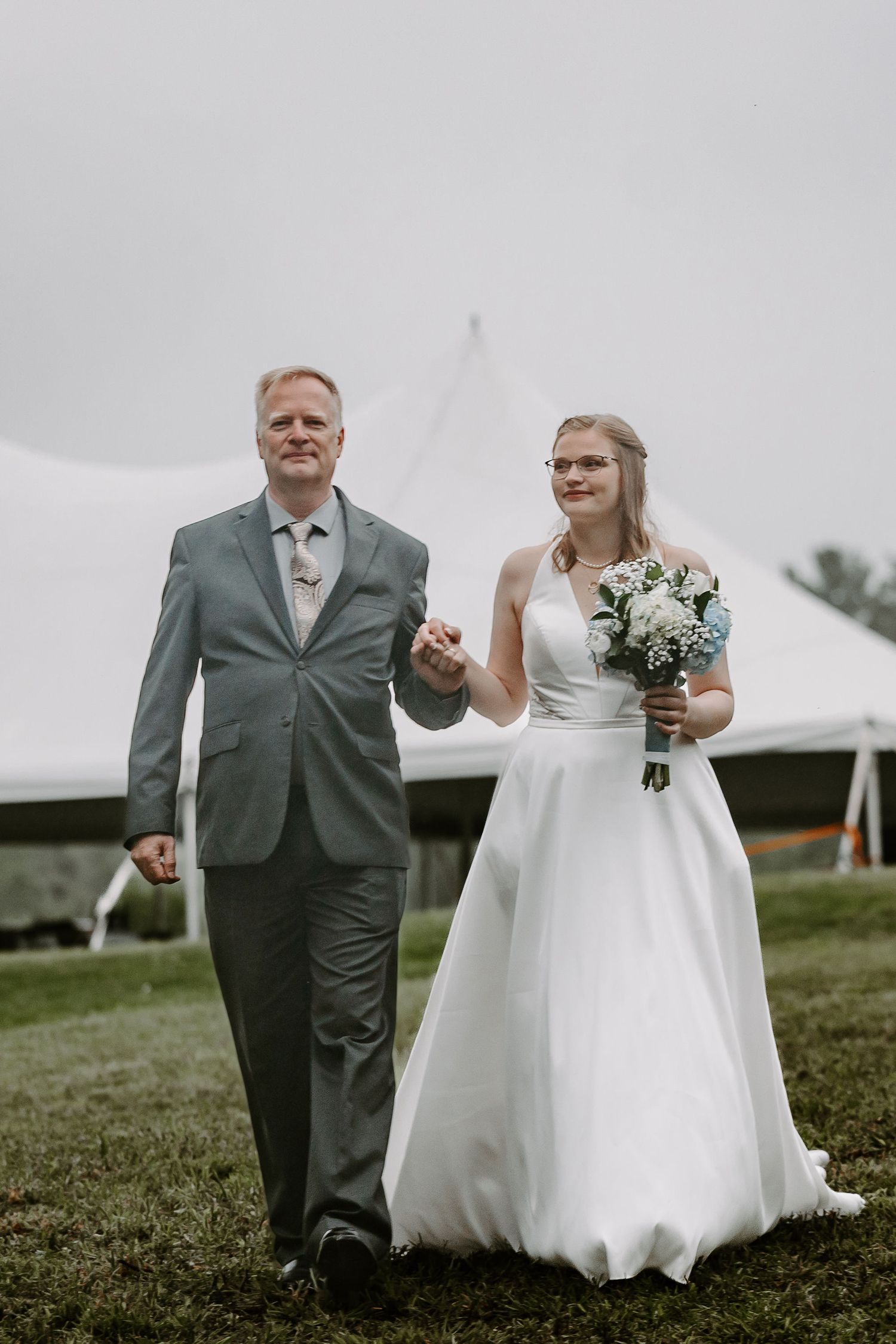 Father of the bride walking daughter down the aisle in white wedding gown at outdoor tent wedding.