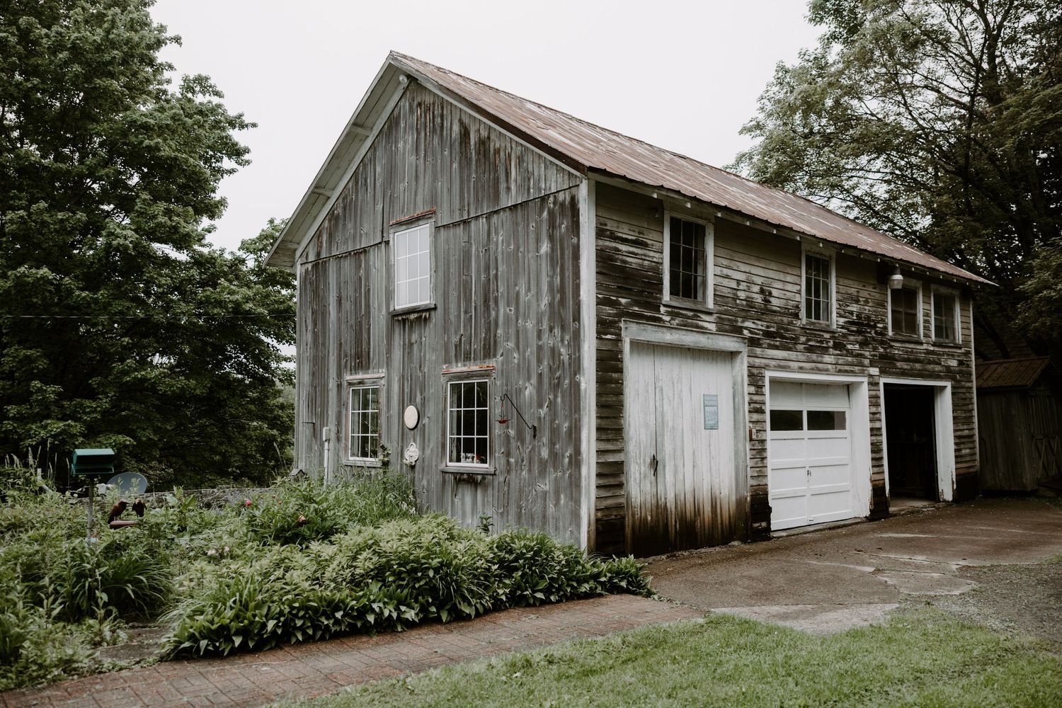 Rustic weathered wooden barn with white garage doors surrounded by green trees.