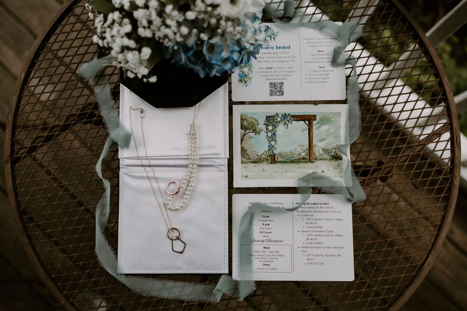 Wedding flat lay with invitation suite, jewelry, blue ribbon, and floral arrangement on wicker table.