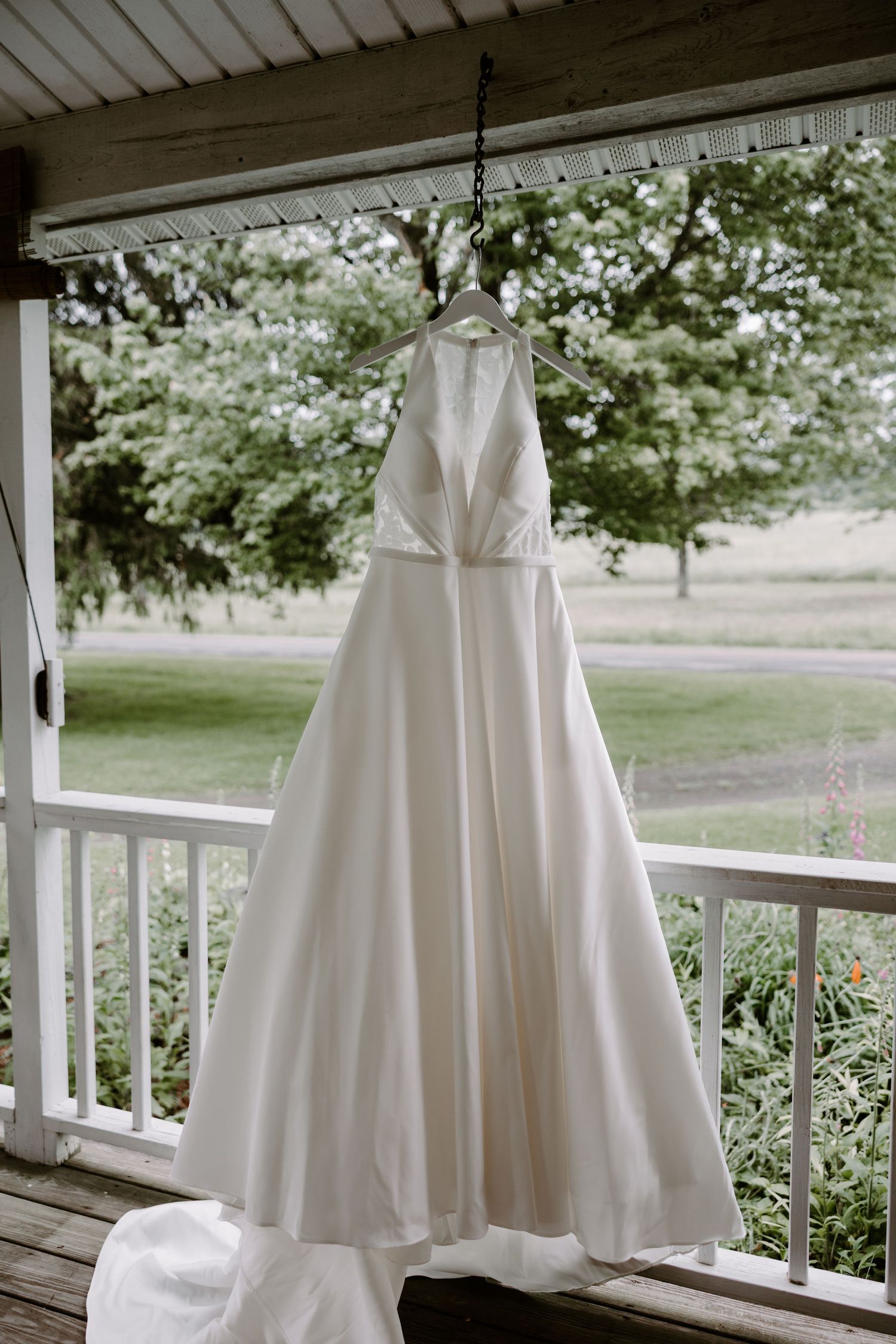 Elegant white halter neck wedding gown hanging on porch with scenic green garden background.