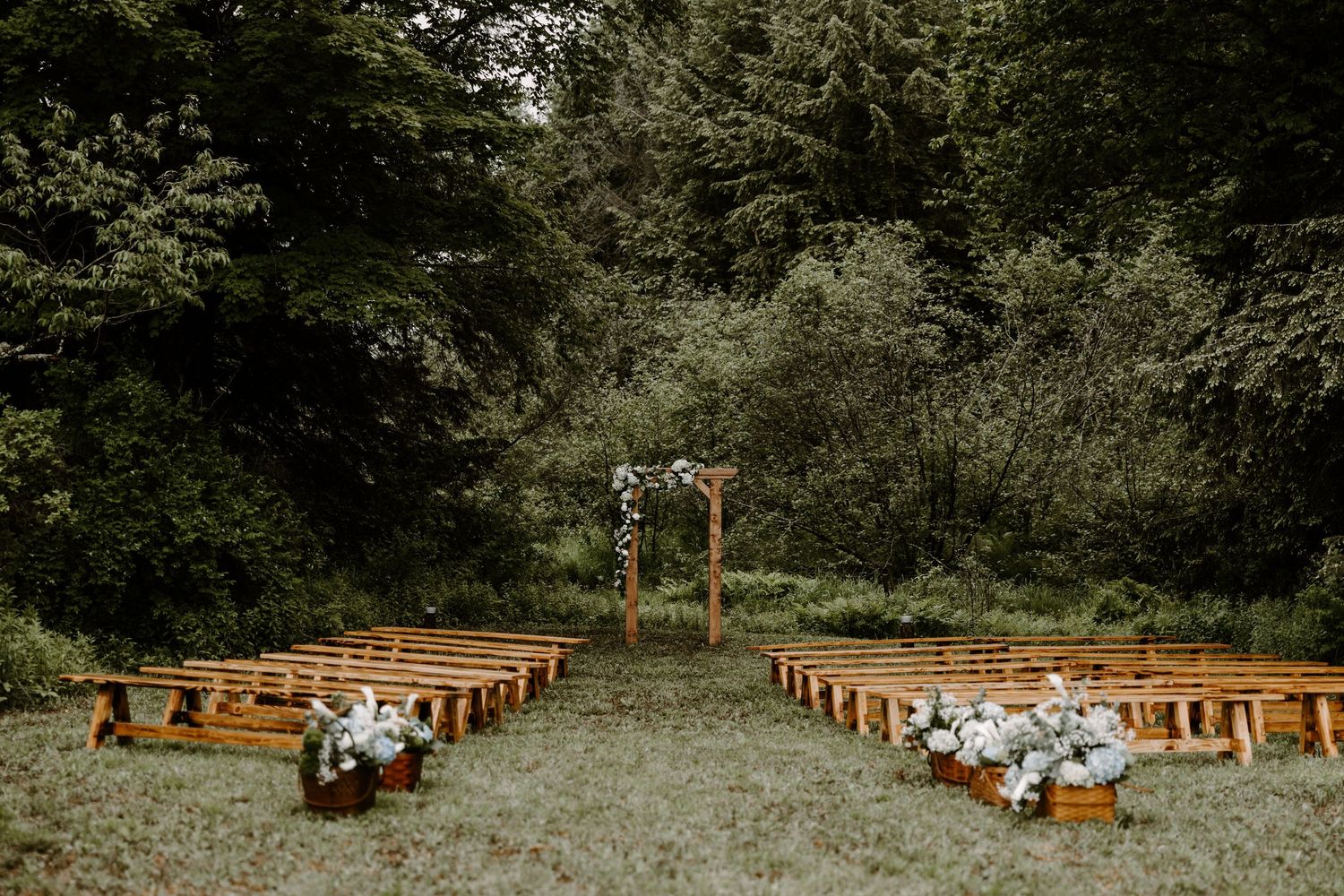 Rustic outdoor wedding ceremony setup with wooden benches, floral arch, and lush green forest backdrop.