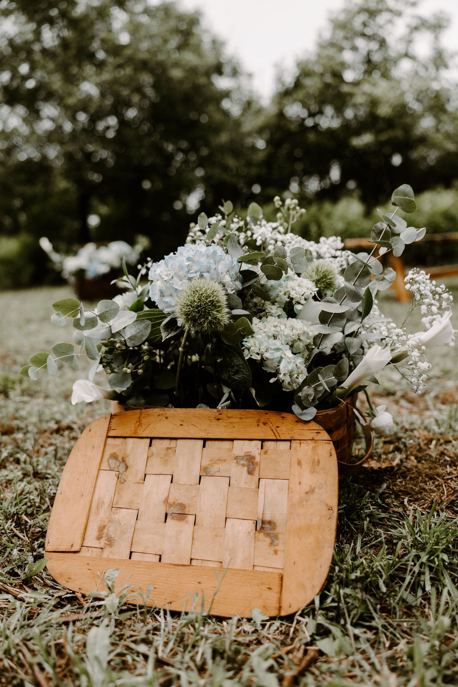 Rustic wooden basket filled with blue hydrangeas and eucalyptus floral arrangement at outdoor wedding.