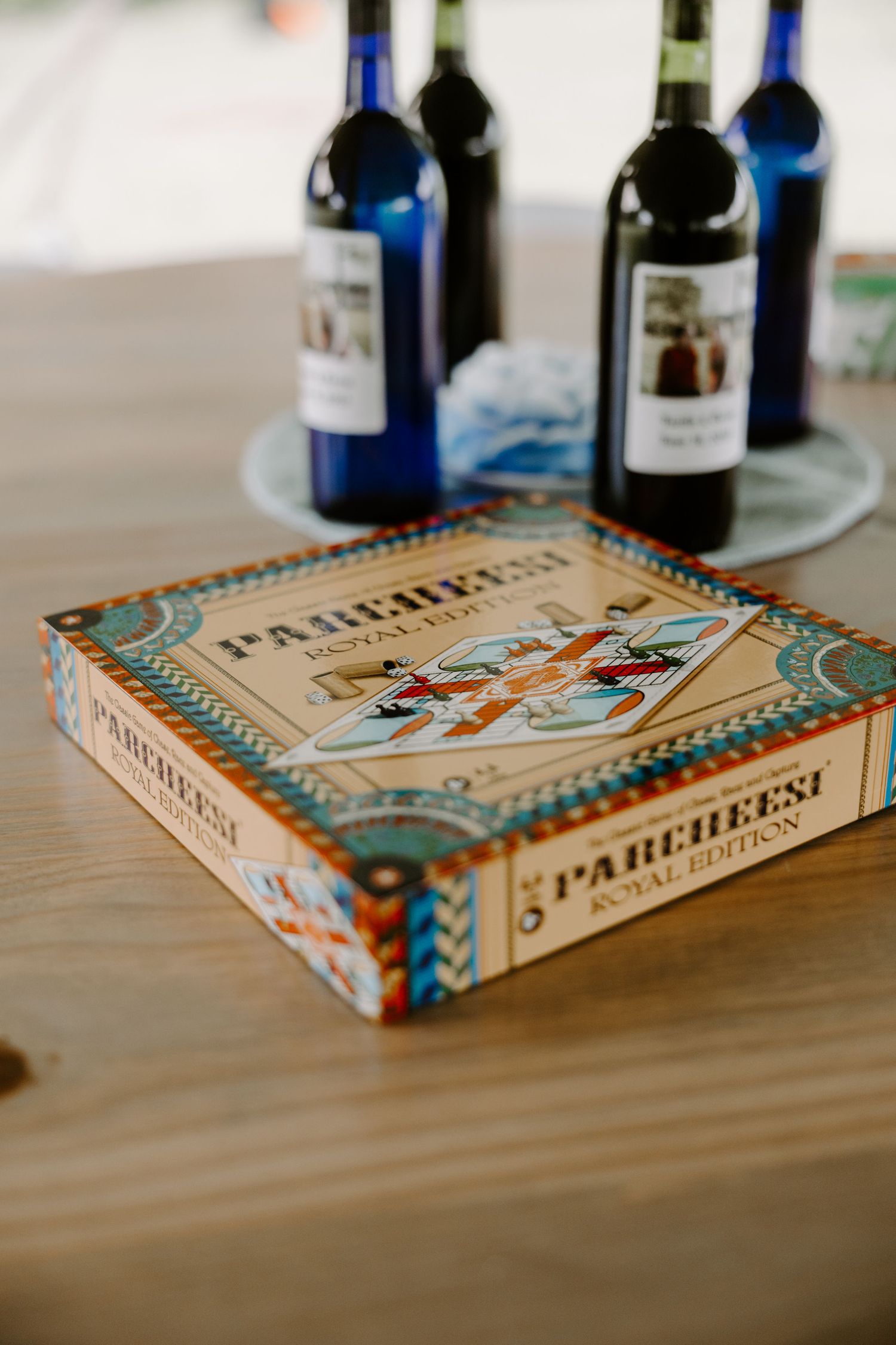 Parcheesi Royal Edition board game box on wooden table with wine bottles in background.