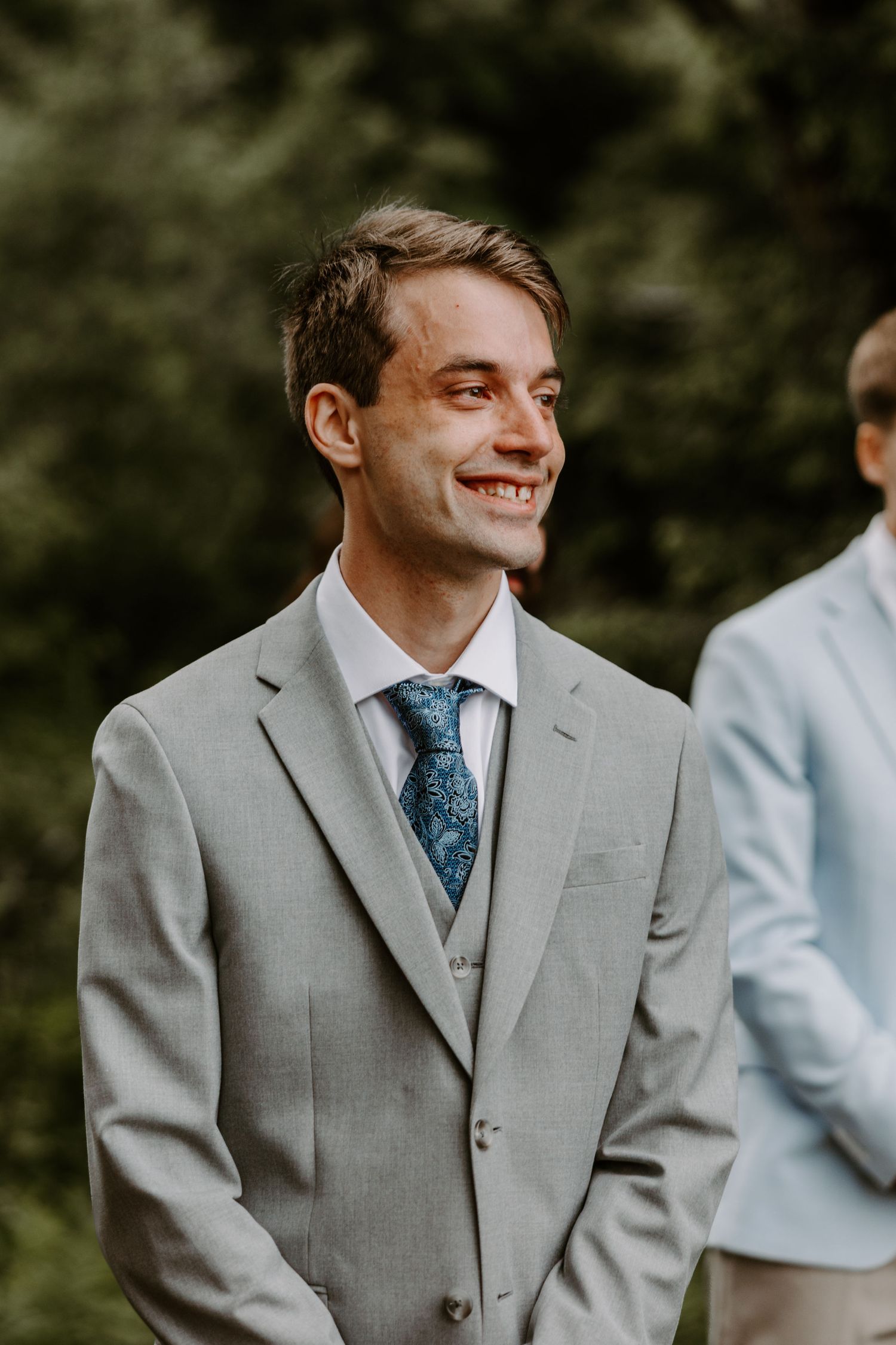 Handsome groom smiling joyfully in gray suit and blue floral tie at outdoor wedding ceremony.
