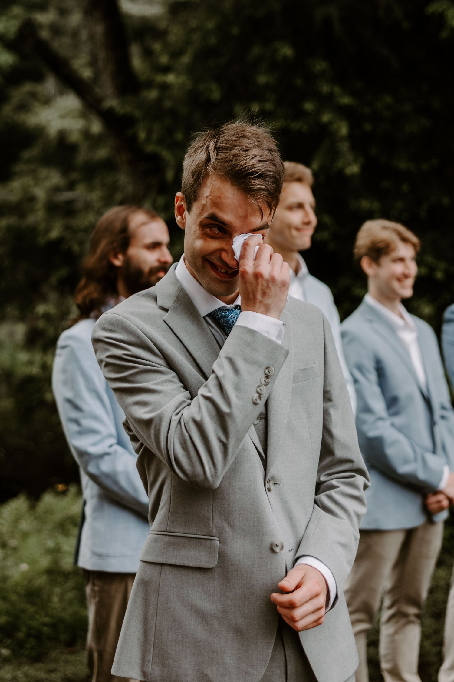 Emotional groom wiping tears during outdoor wedding ceremony with groomsmen in gray suits behind him.