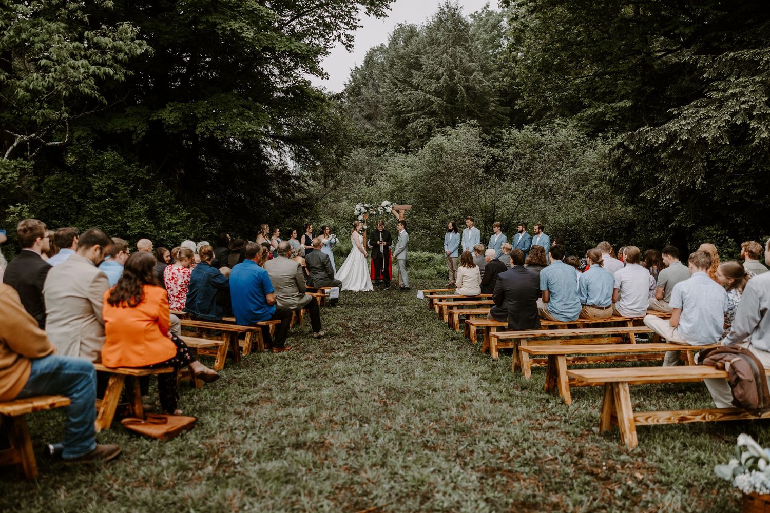 Guests seated on wooden benches at elegant outdoor woodland wedding ceremony.