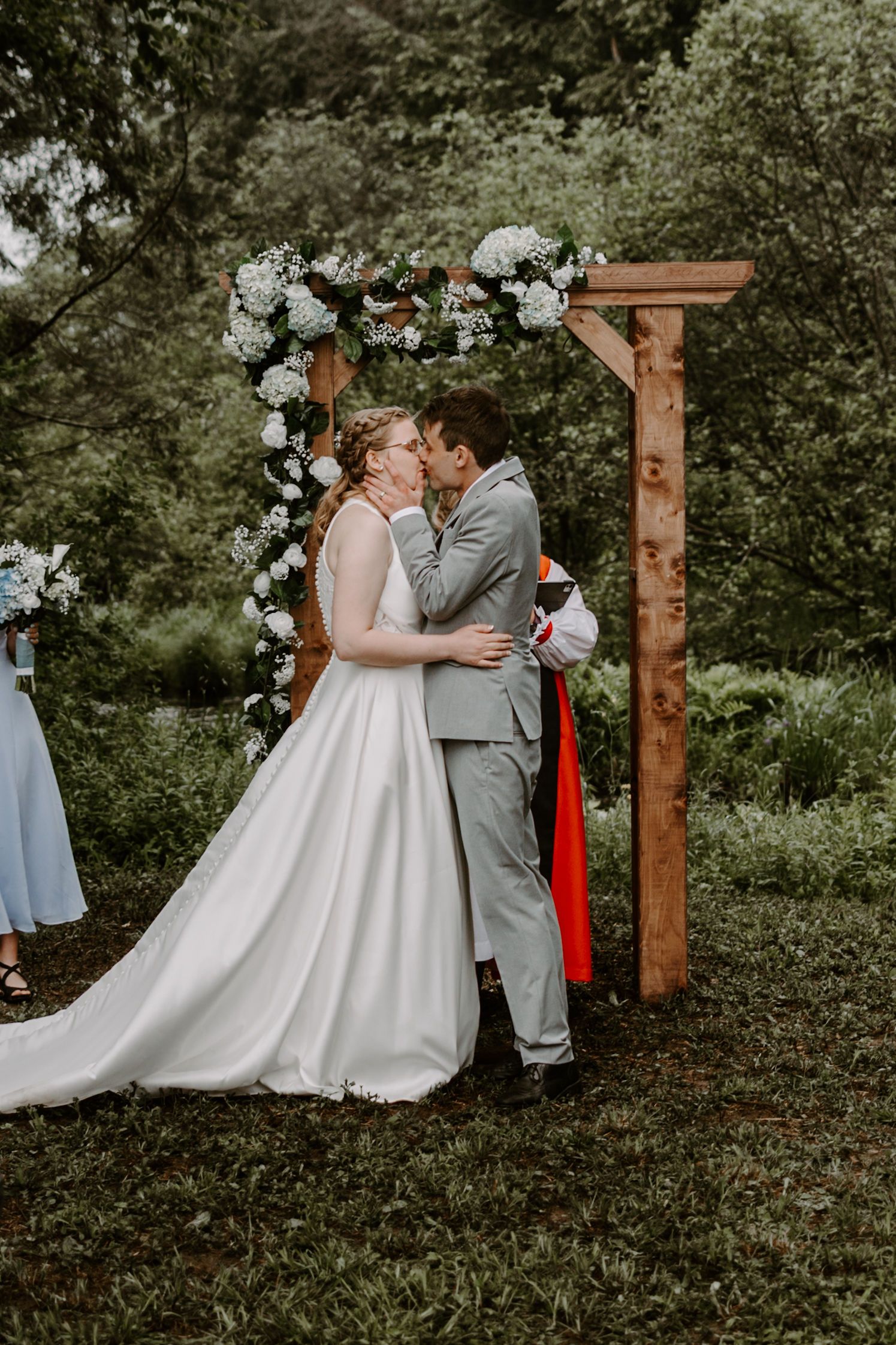 Bride and groom sharing first kiss under floral wooden arch at outdoor ceremony.