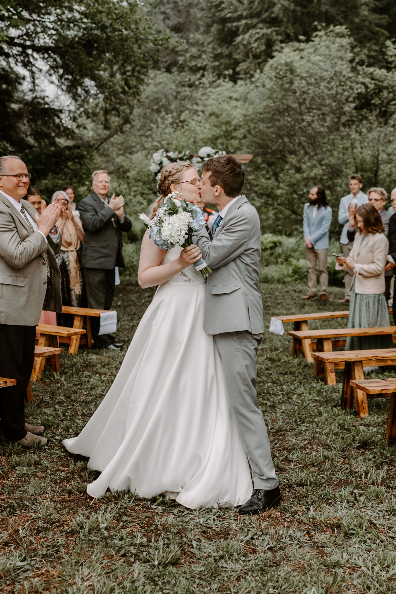 Newlyweds kissing in aisle as guests applaud at outdoor woodland wedding ceremony.