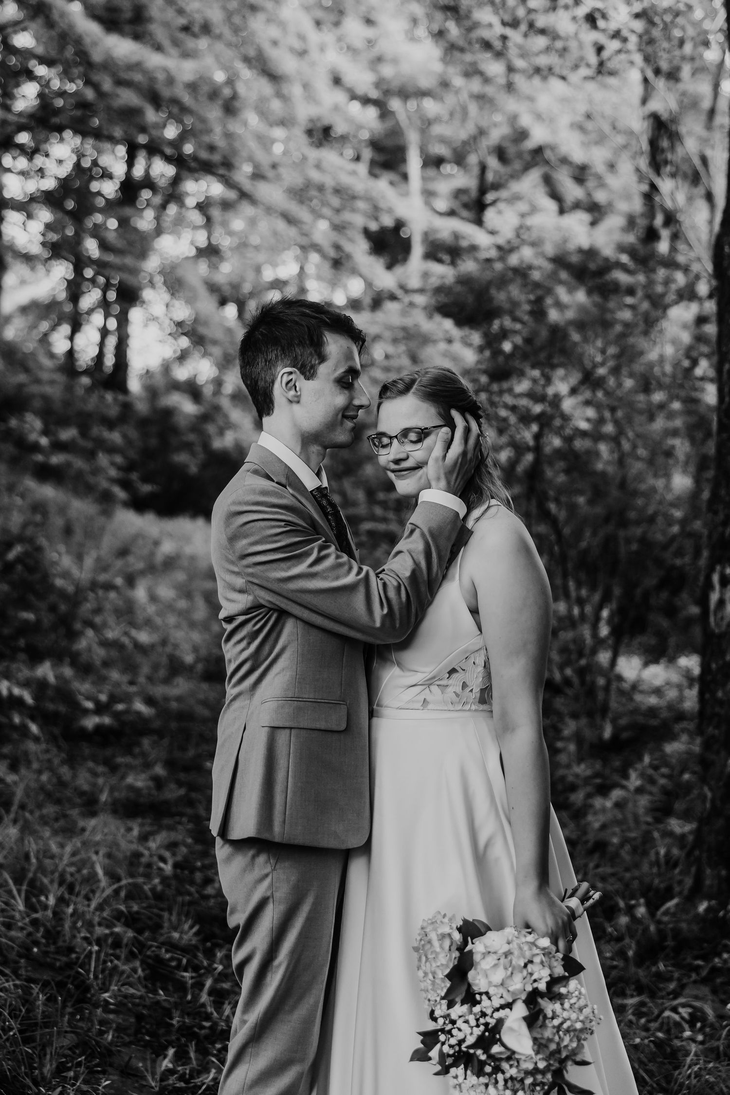 Black and white portrait of groom tenderly touching brides face in a lush forest setting.