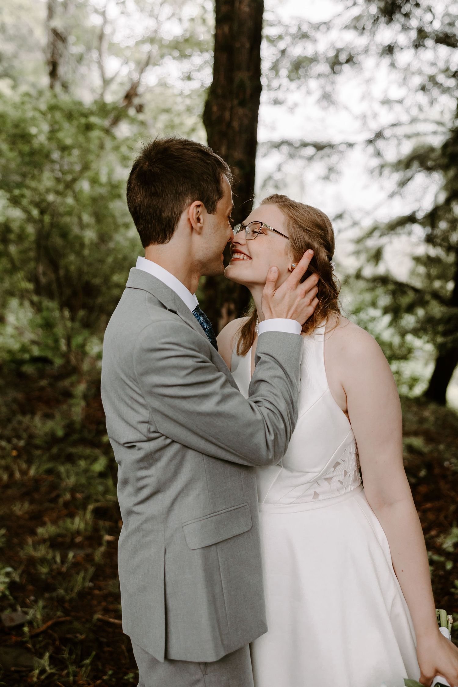 Groom gently holding brides face as she smiles up at him surrounded by green forest trees.