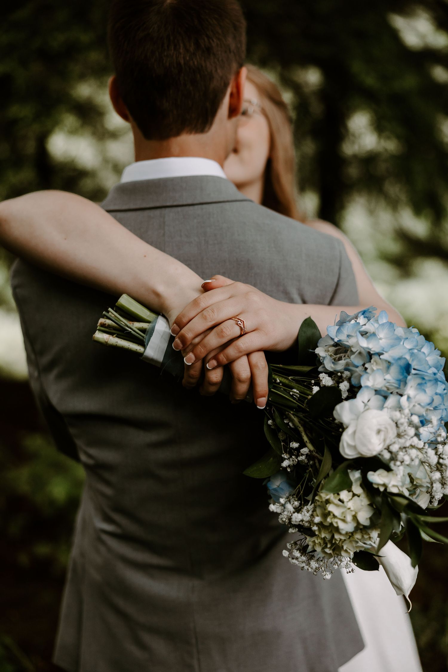 Bride holding blue hydrangea bouquet while embracing groom in gray suit at outdoor wedding.