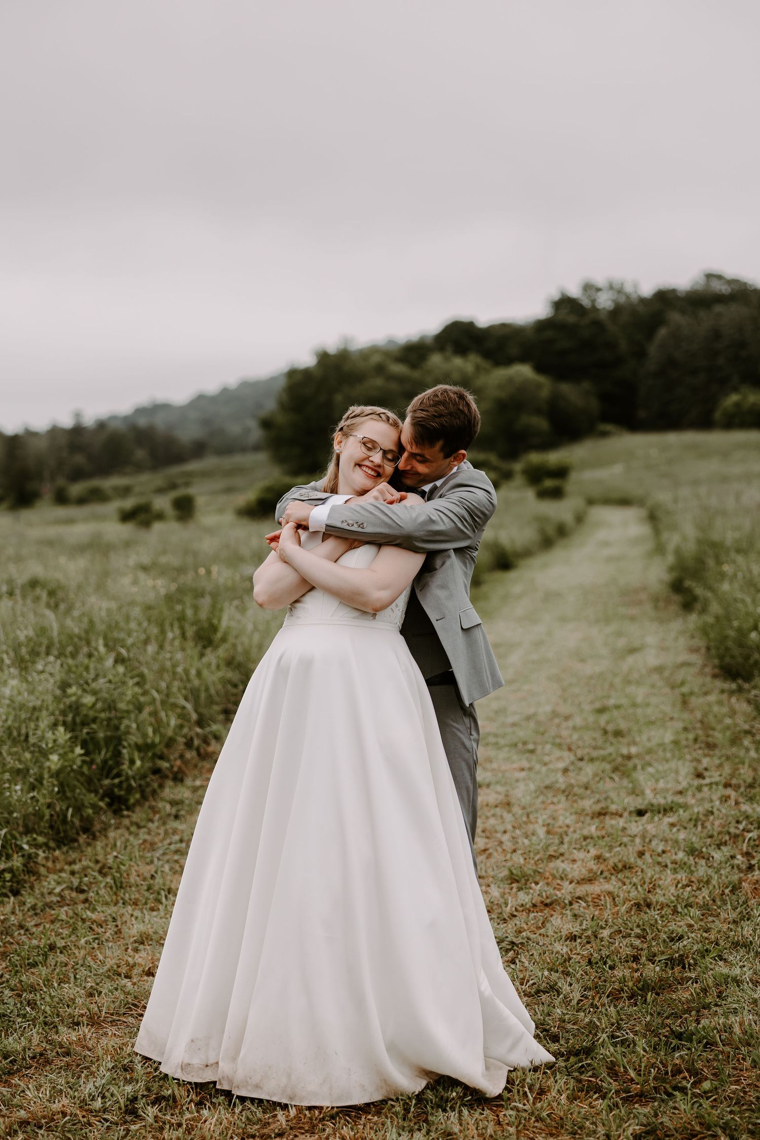 Groom embracing bride from behind in lush green meadow during romantic outdoor wedding portrait session.