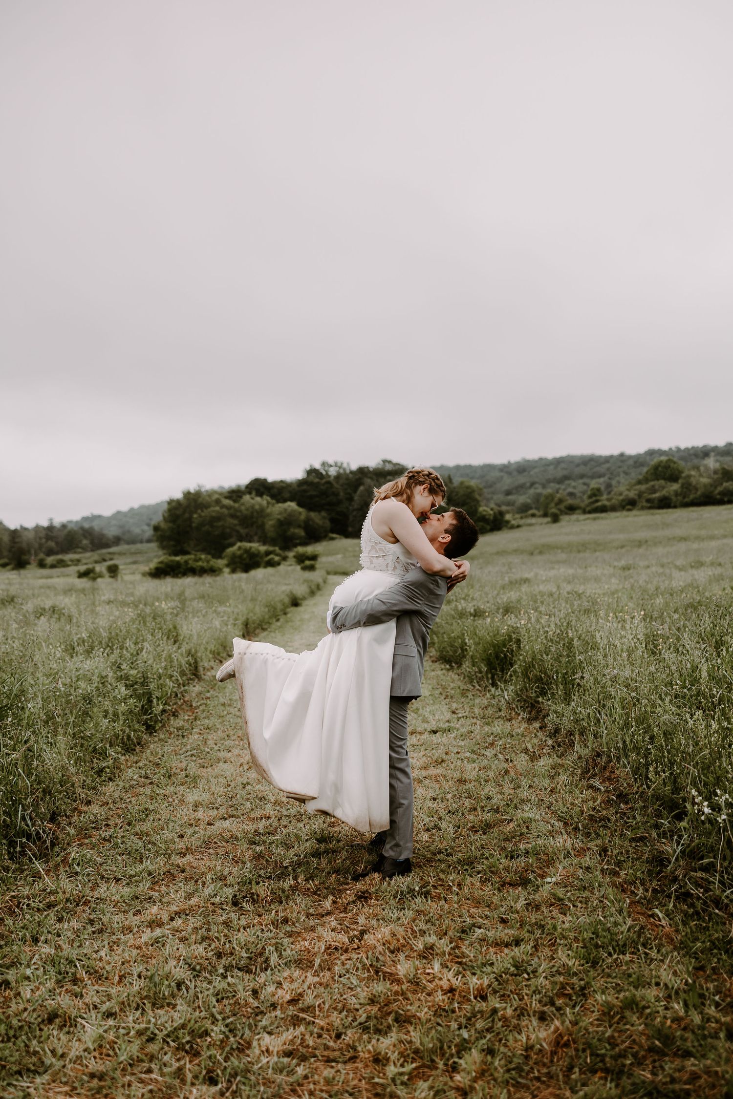 Groom lifting bride in white gown in open green field during romantic outdoor wedding photo session.