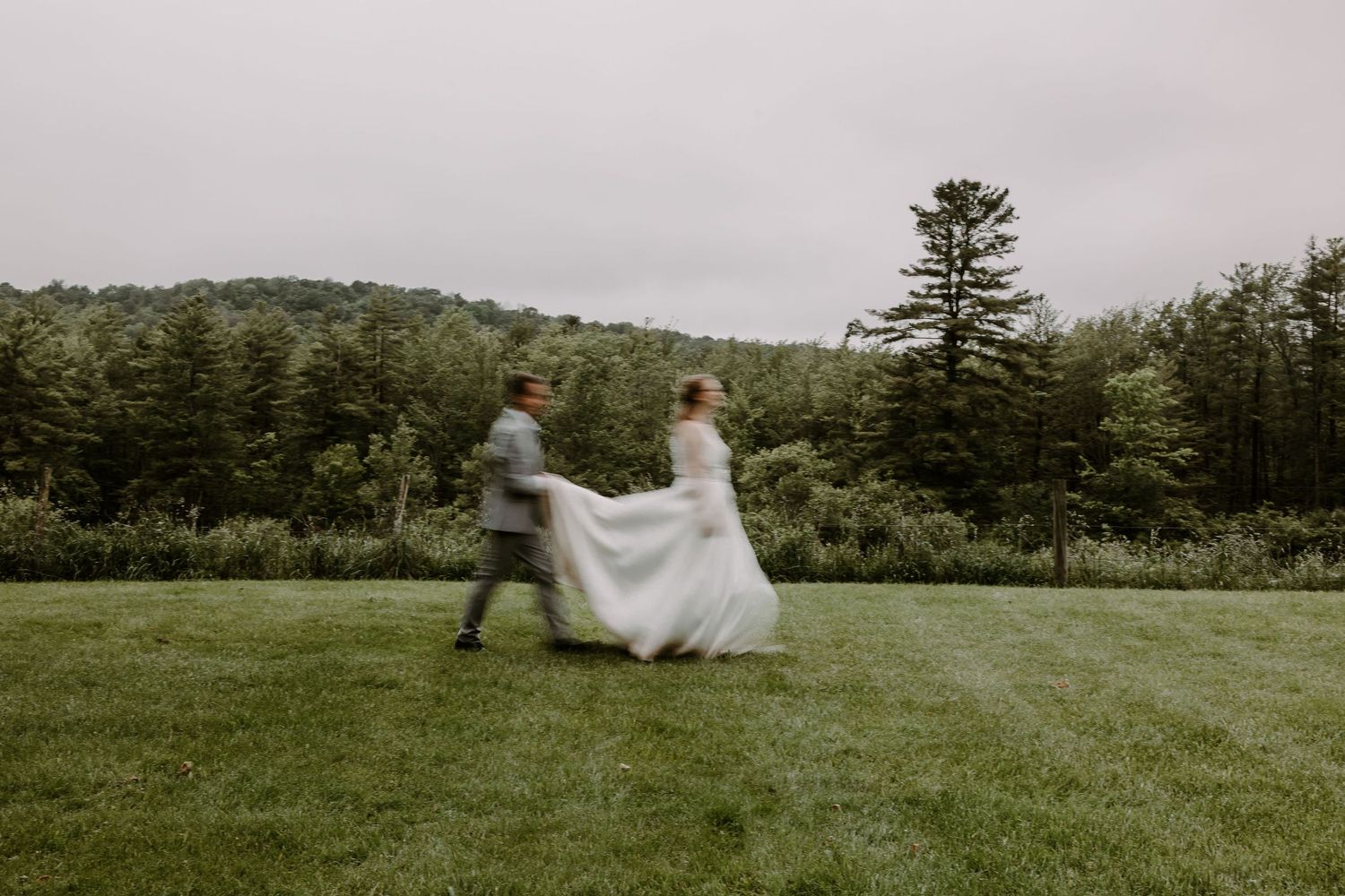 Bride and groom running playfully across green lawn with forested hills behind.