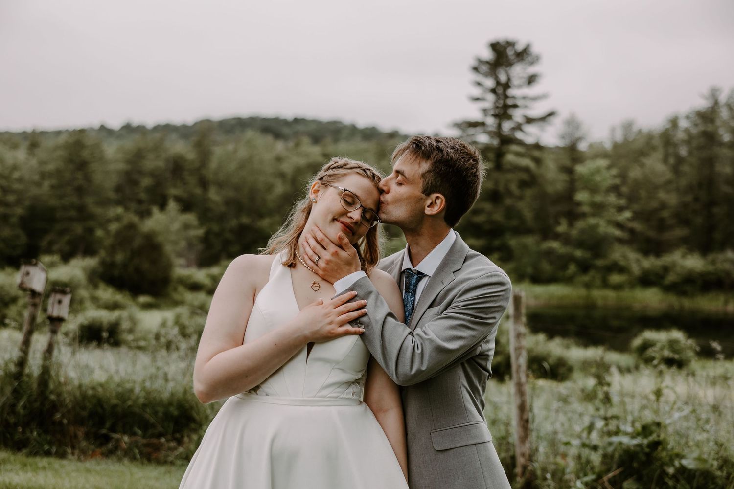 Groom tenderly kissing bride on cheek in green countryside setting after outdoor wedding ceremony.