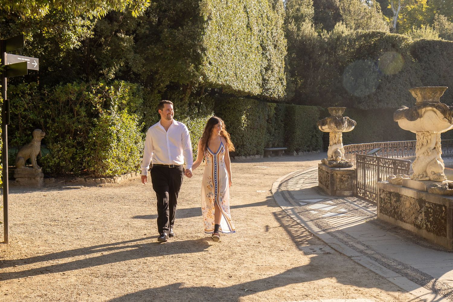 A couple walks hand in hand along a sunlit stone path with ornate garden features in the background.