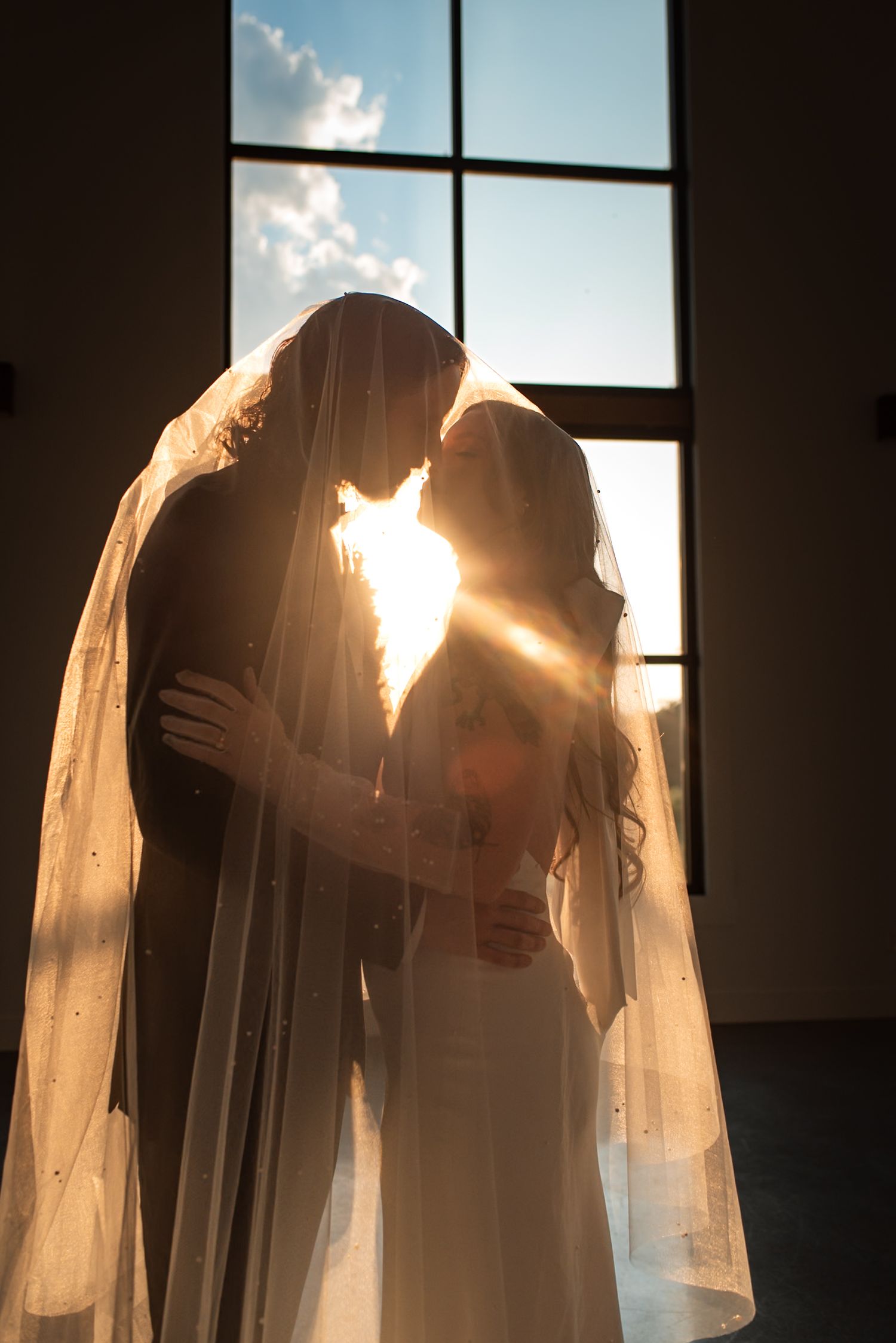 Silhouetted couple in wedding attire share an intimate moment backlit by sunlight streaming through a window.