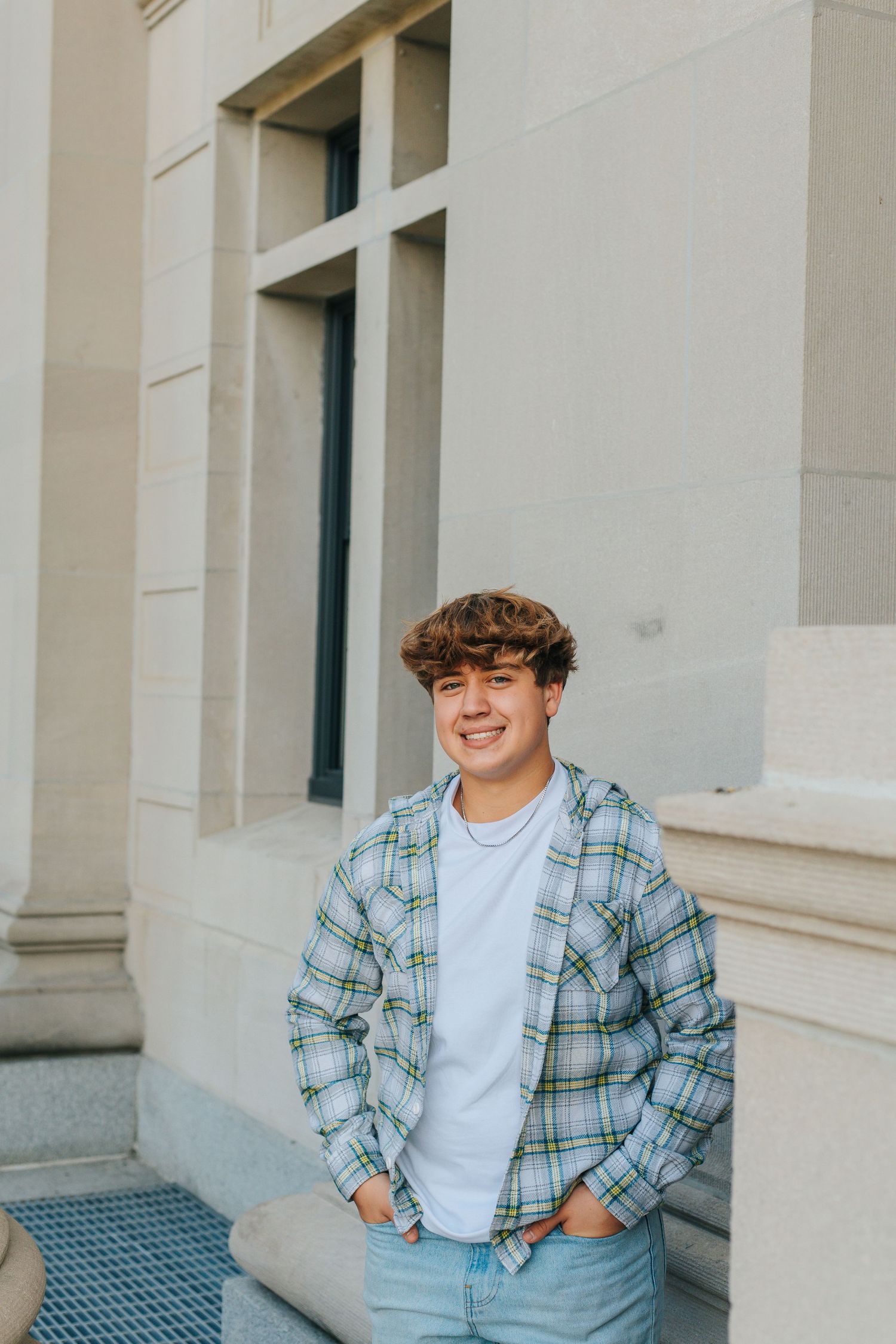 A person in a plaid shirt and white t-shirt sits outdoors against a light-colored building wall.
