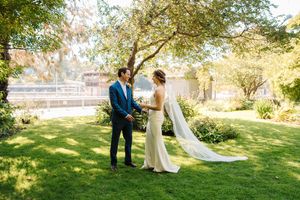 A couple in wedding attire stands together on a sunlit lawn with trees and a building in the background.