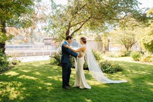 A couple shares a romantic moment during their outdoor wedding ceremony on a sunlit lawn surrounded by trees.