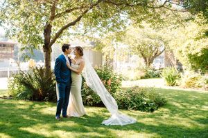 A couple in wedding attire embraces in a sunlit garden setting with a flowing white veil and lush greenery.