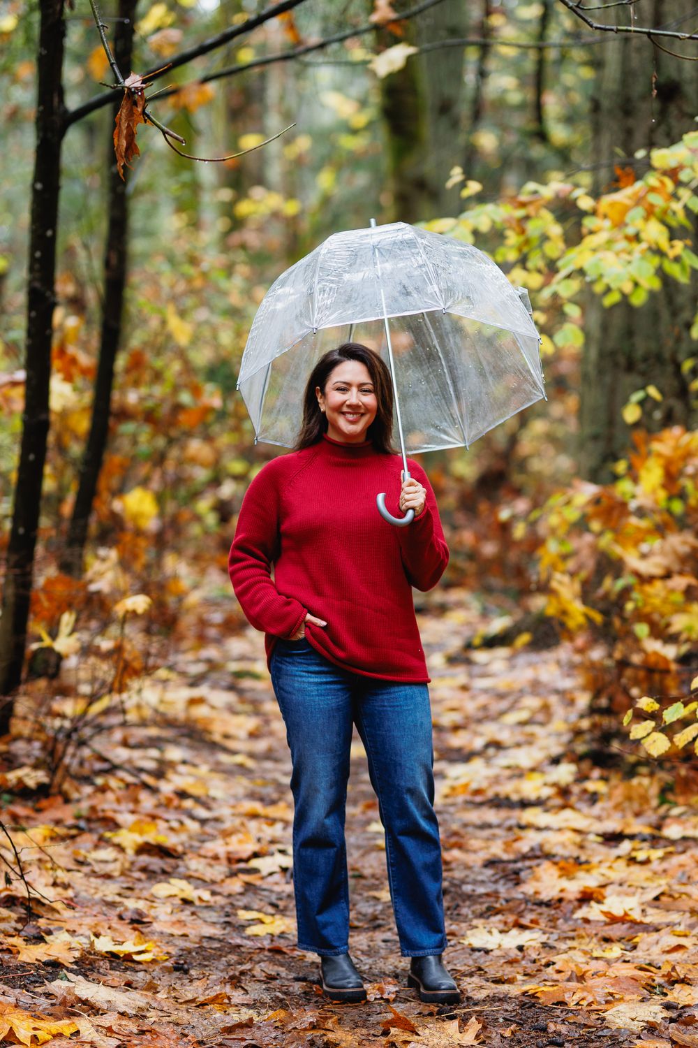Happy woman in red sweater and jeans walking with a transparent umbrella in fall forest.