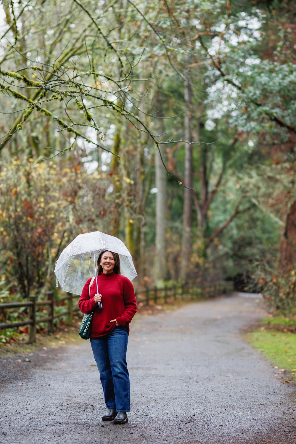 Smiling woman in red sweater carrying a clear umbrella while strolling a rainy forest path.