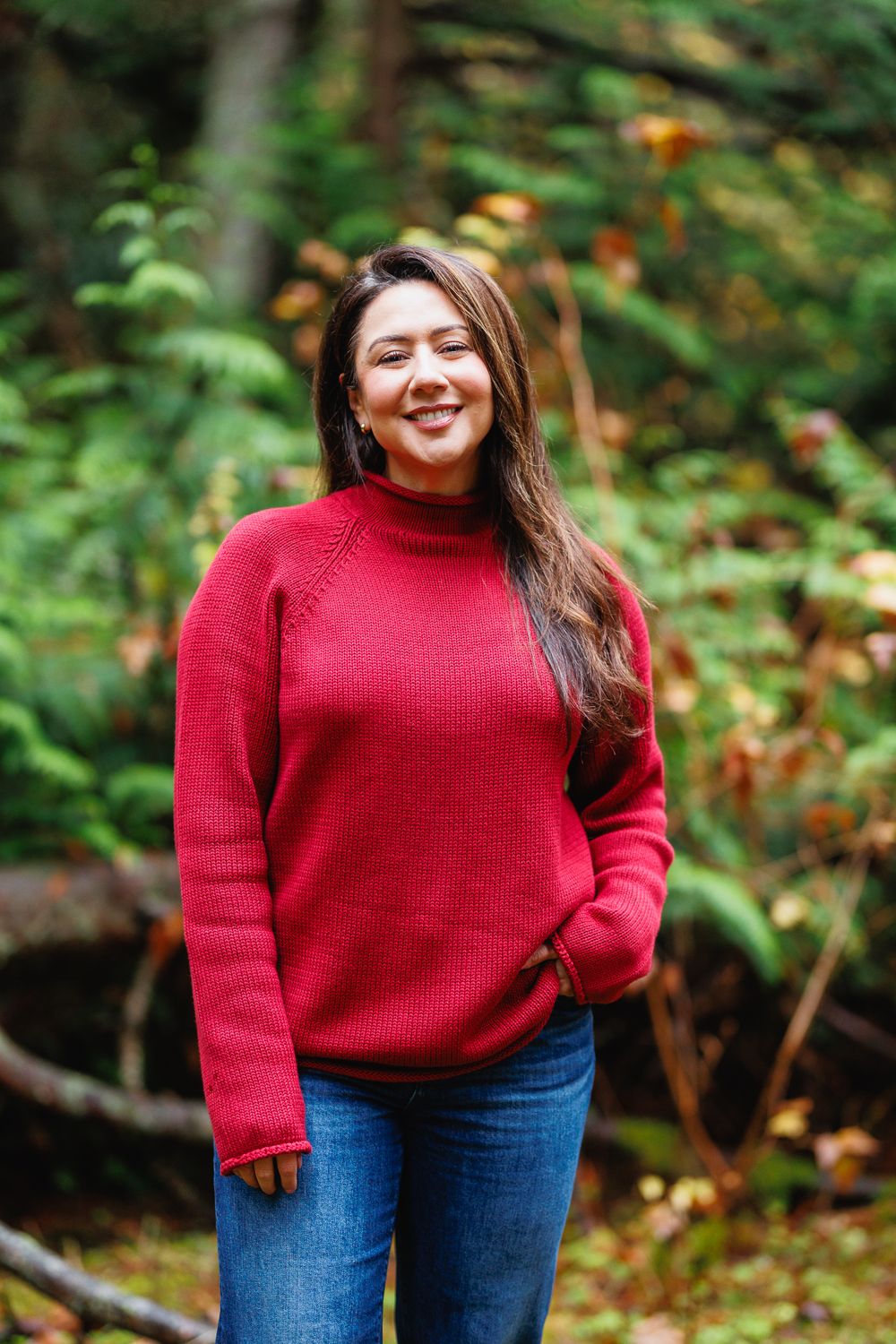Joyful woman in red sweater standing and smiling in a lush green forest setting.