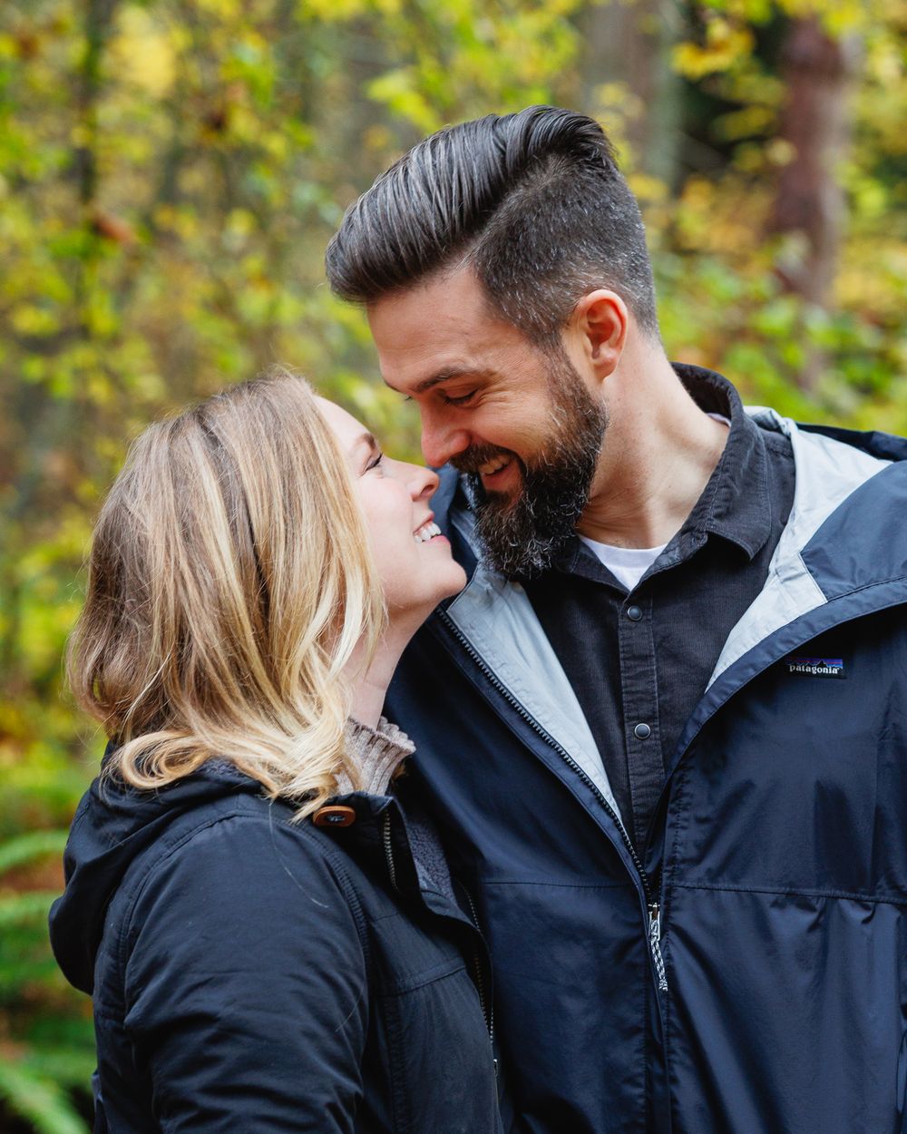 Couple smiling and nearly kissing outdoors in autumn forest setting.