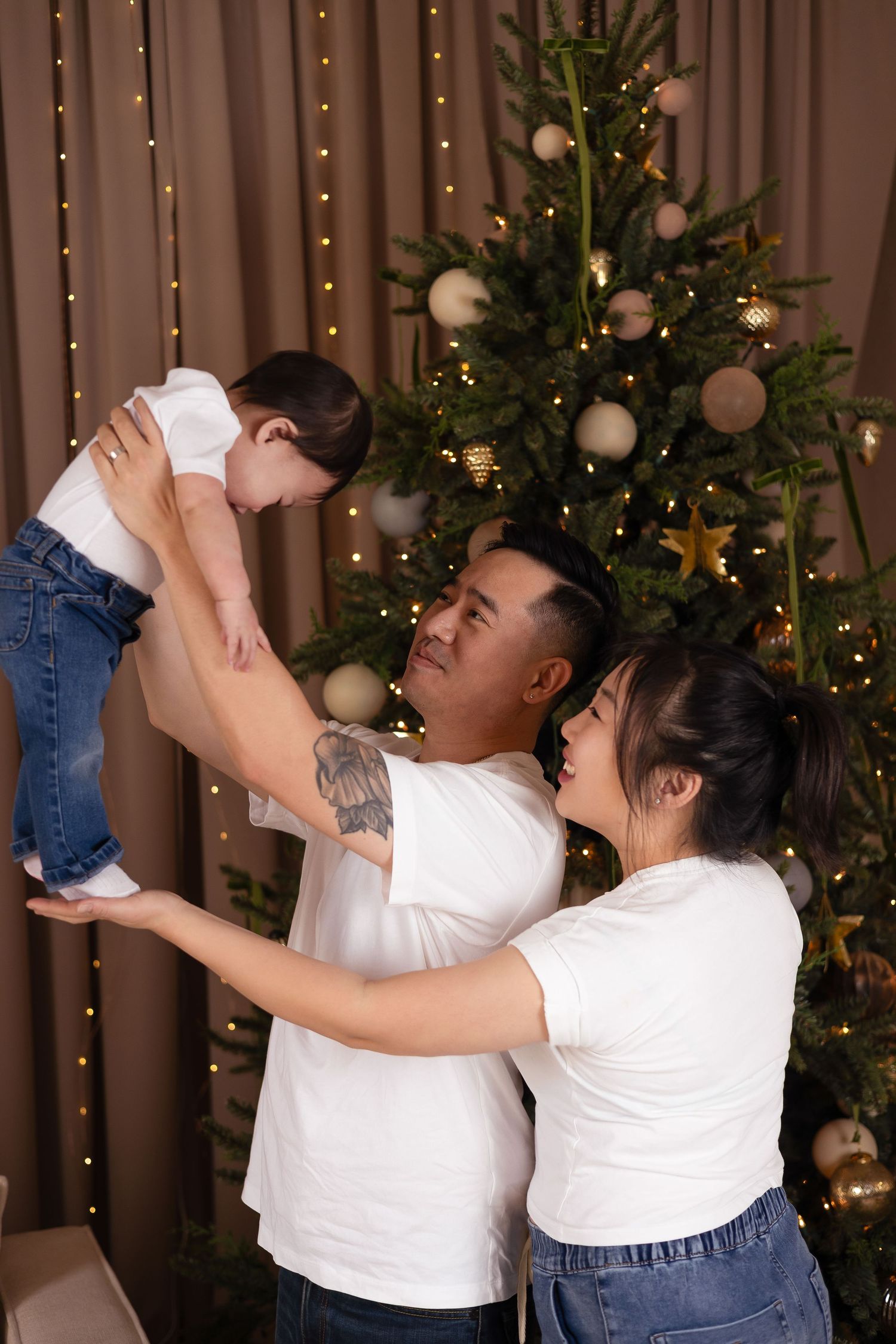 Asian family in matching white tees joyfully lifts baby near a decorated Christmas tree.