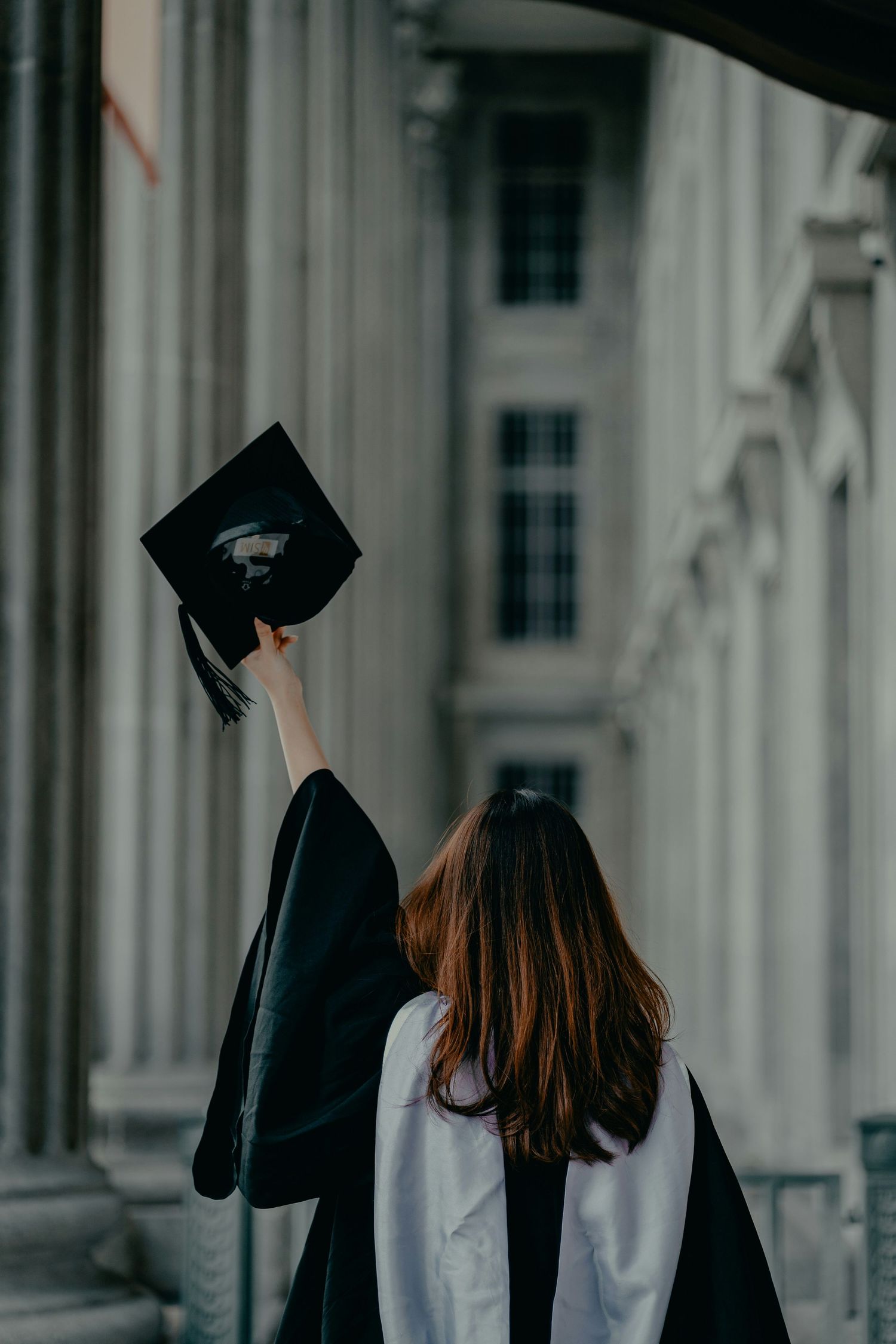 Graduates in black caps and gowns celebrate by throwing their mortarboards in the air against a white building backdrop.