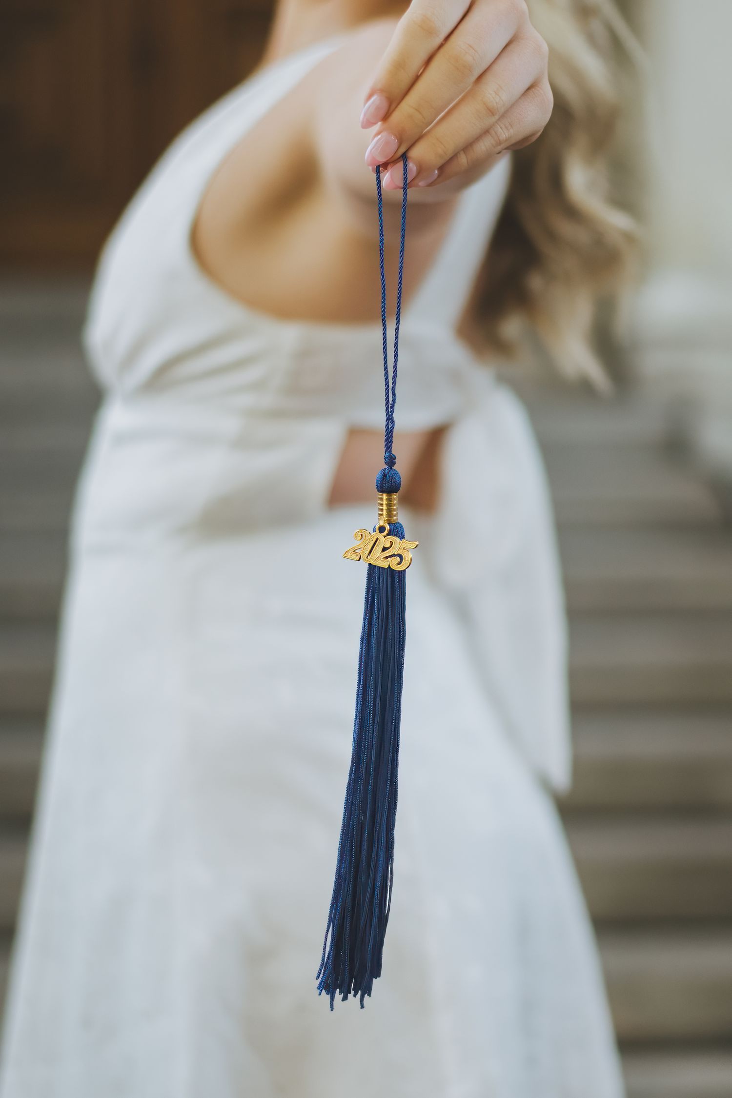 Someone in a white dress holds a navy blue graduation tassel with gold embellishment against a blurred background.
