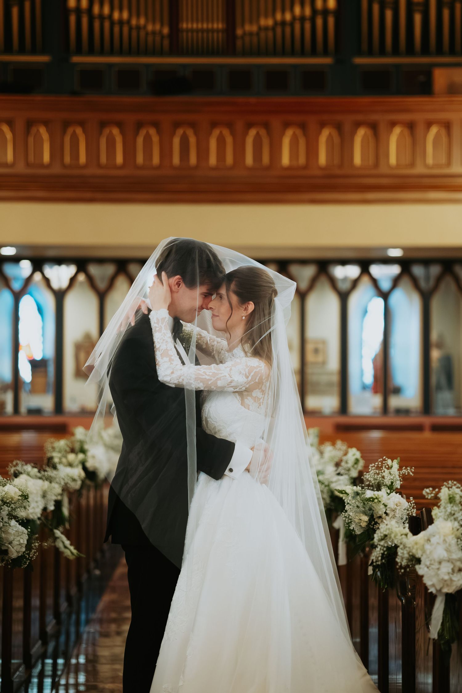 Bride and groom embrace under a sheer veil in a church aisle lined with white floral arrangements and wooden pews.