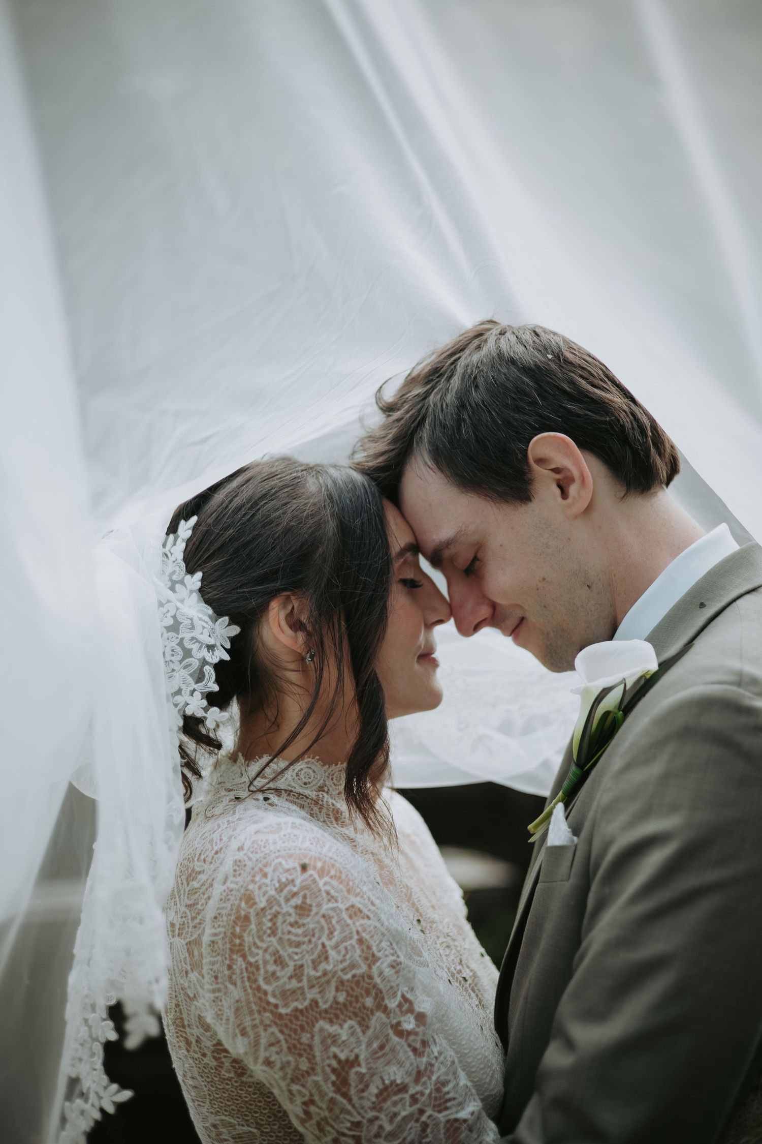 Bride and groom touch foreheads with eyes closed beneath a sheer white veil, her lace gown and his gray suit with boutonniere.