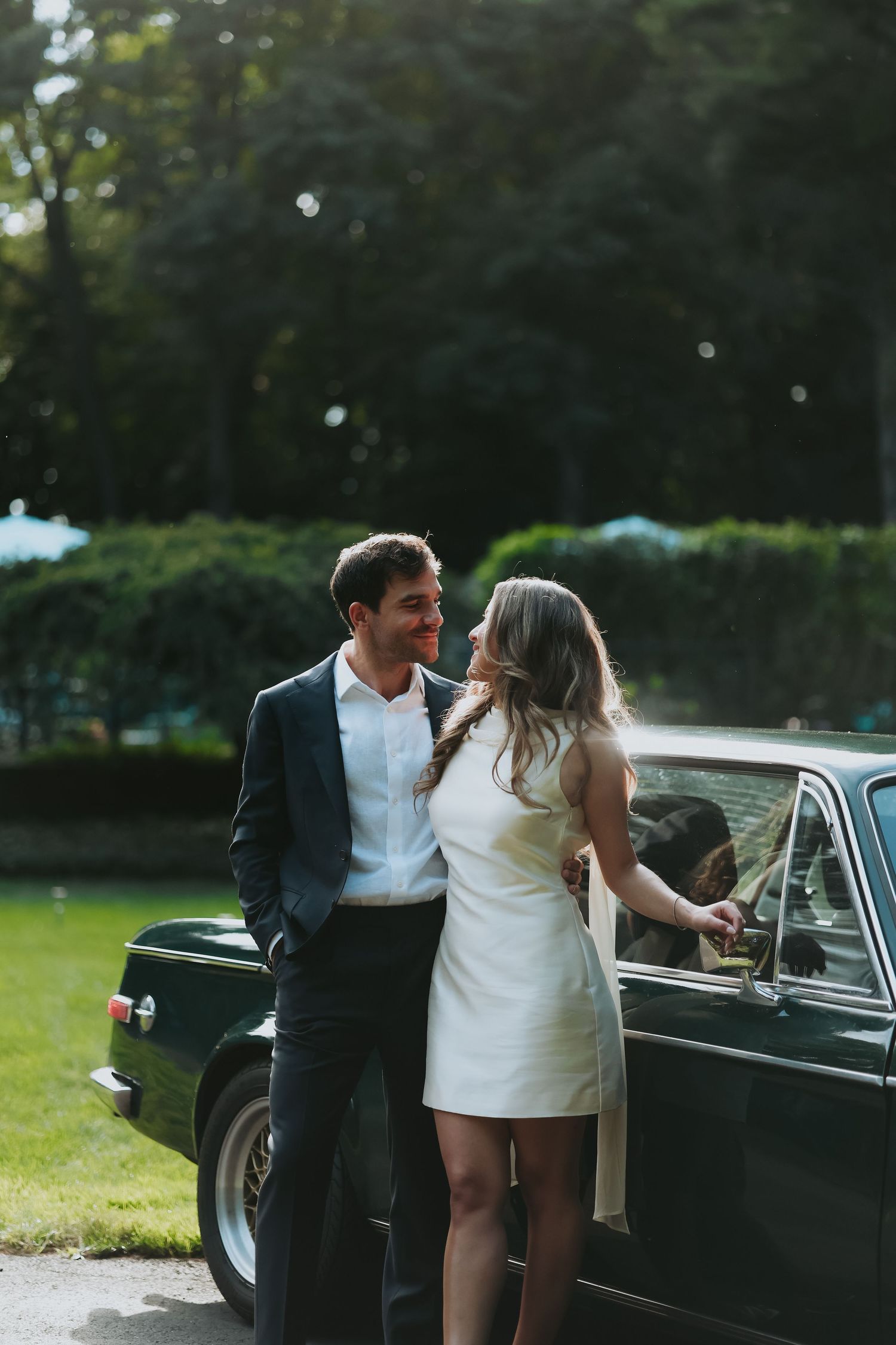 Smiling couple in formal attire standing close beside a vintage black car on a sunlit lawn surrounded by green trees.