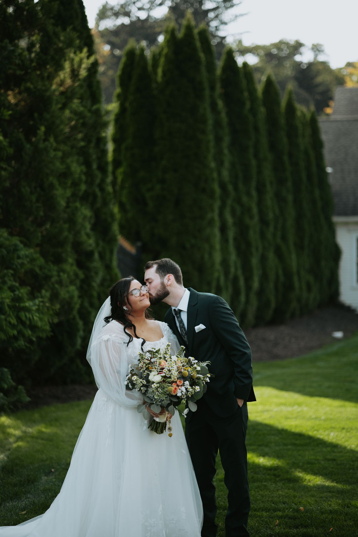 Groom in a dark suit kisses bride in a white veiled gown holding a floral bouquet beside tall evergreen trees on a sunny lawn.