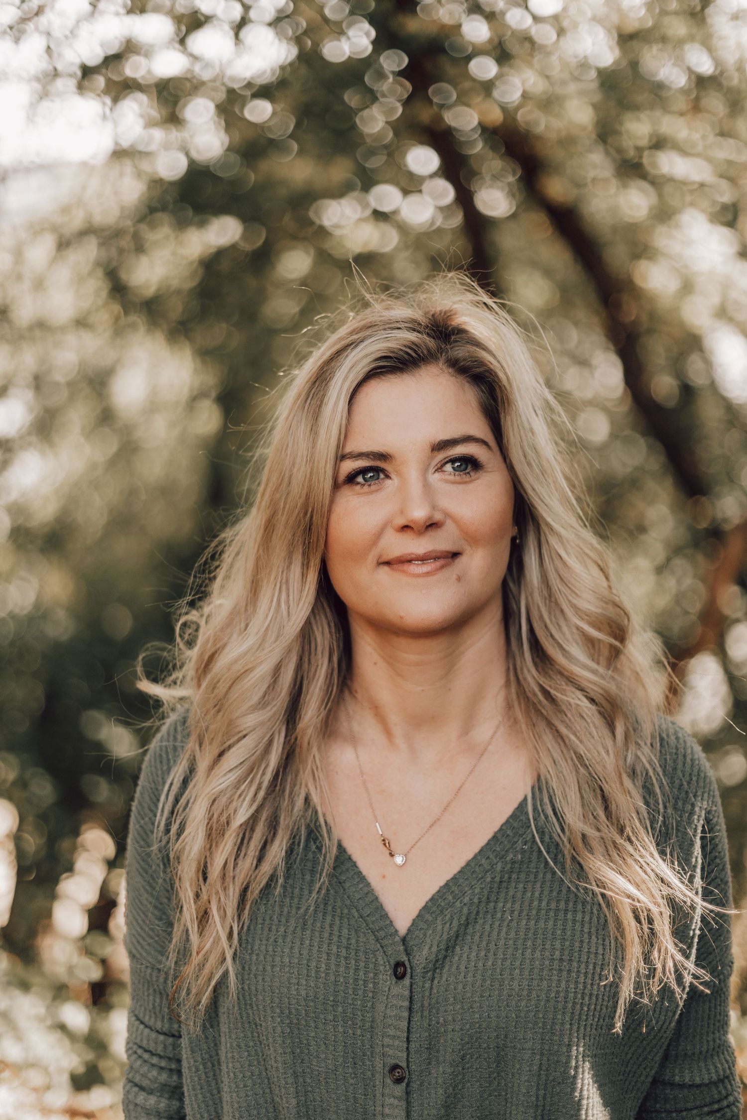 Smiling blonde woman in green cardigan with bokeh tree background in warm outdoor portrait.