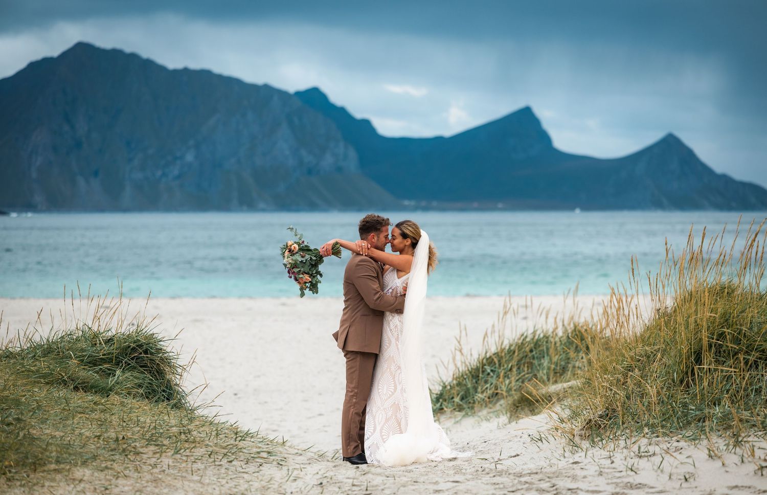 A romantic wedding photoshoot on a scenic beach with dramatic mountain peaks and blue skies in the background.