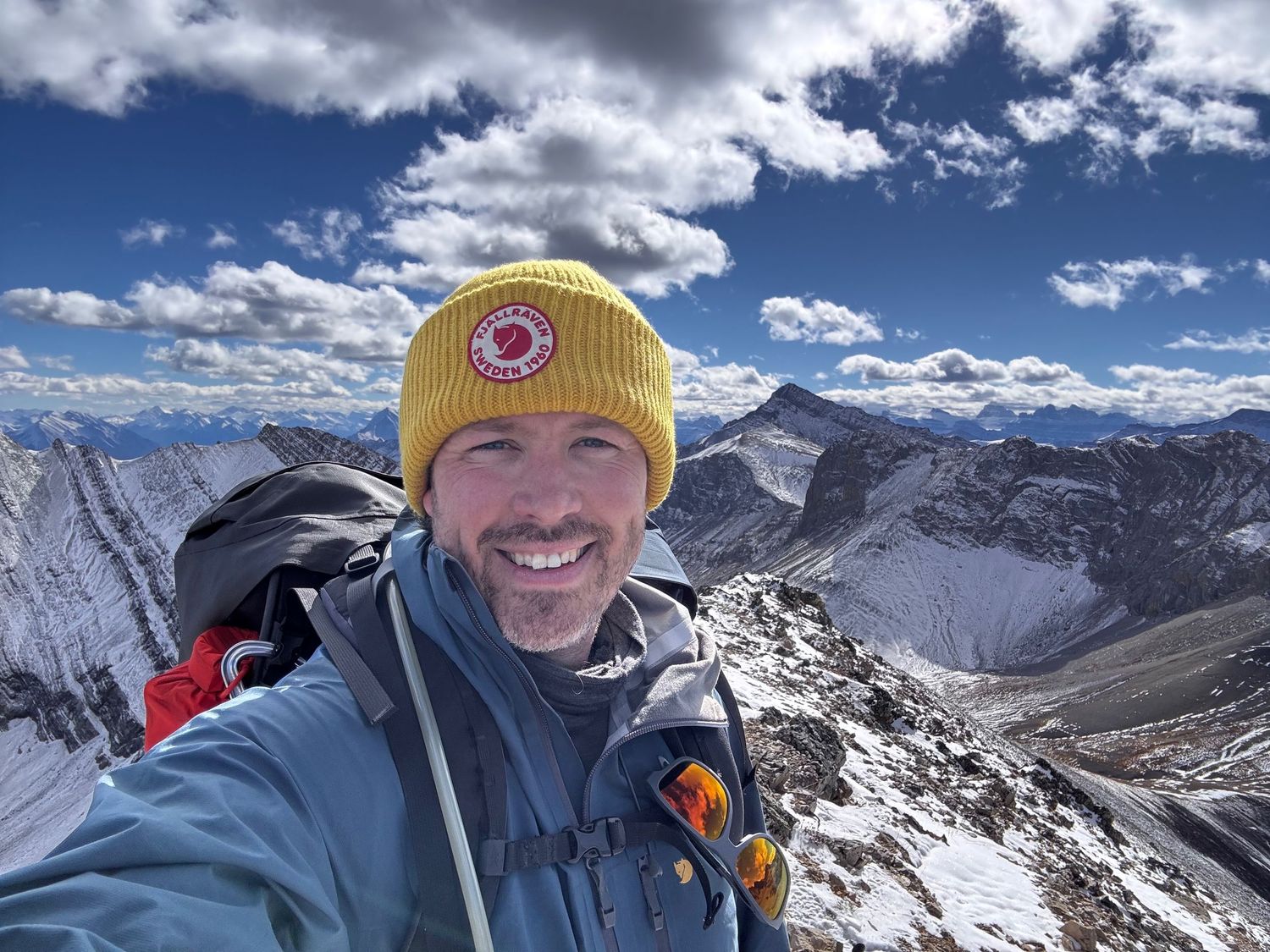 Evan Will - yellow hat stands atop a snowy mountain summit with dramatic peaks and clouds in the background near Nordegg, Alberta. Abraham