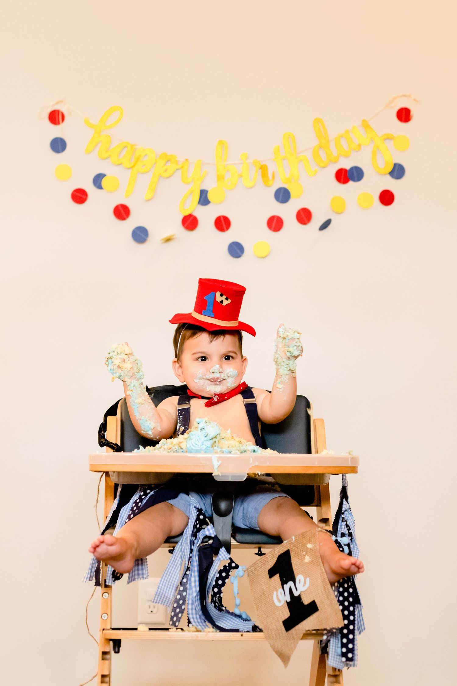 Vintage wooden high chair decorated for first birthday party with red hat and colorful garland backdrop.