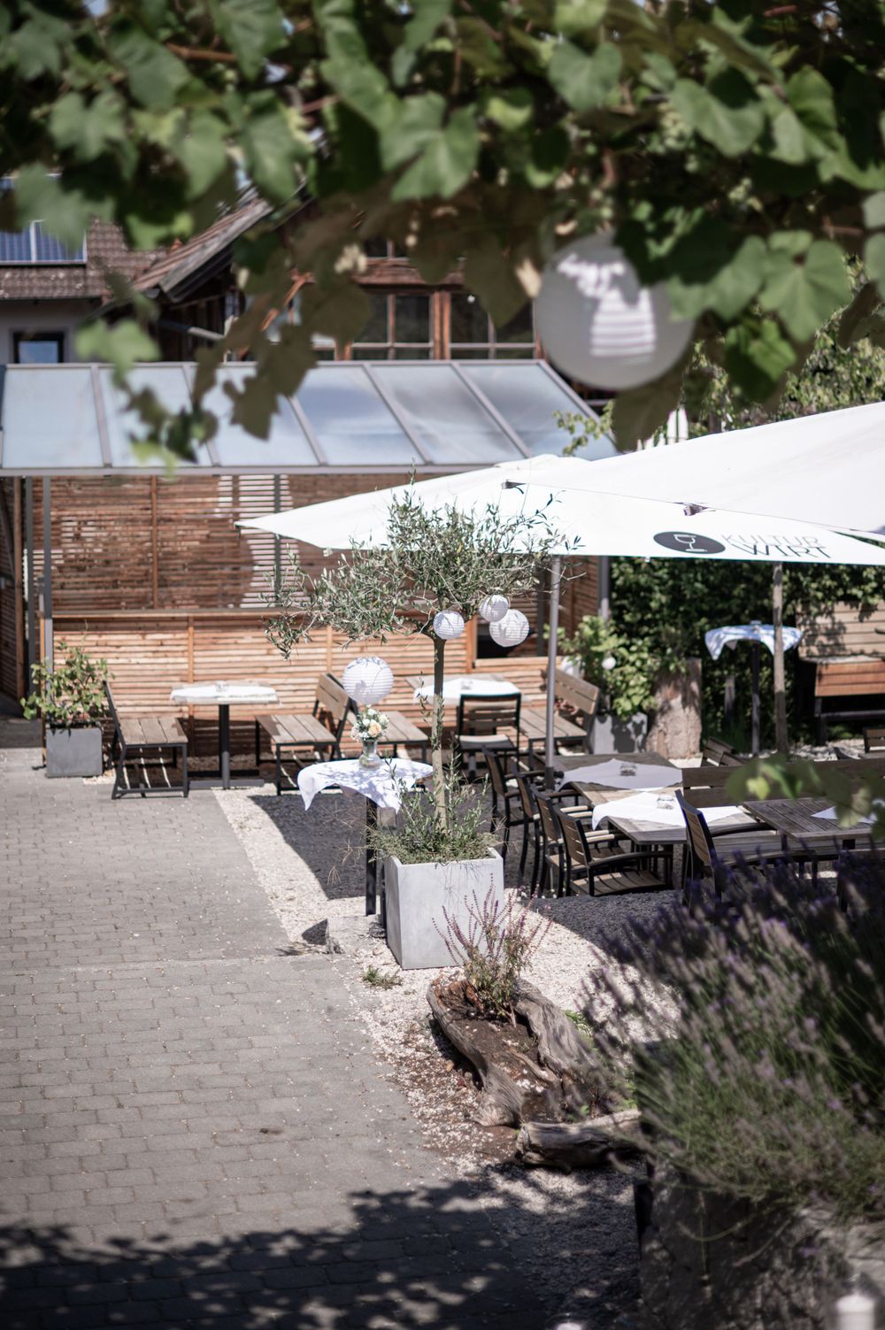 Gemütliche Restaurantterrasse mit Holzmöbeln, Sonnenschirmen und Gartengestaltung bei gedämpftem Sonnenlicht und im Schatten.