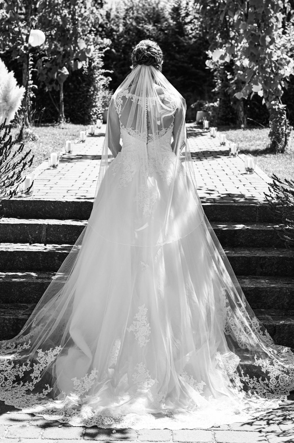 Eine Braut in einem fließenden weißen Hochzeitskleid und Schleier steht auf einer Schwarz-Weiß-Fotografie auf steinernen Stufen.
