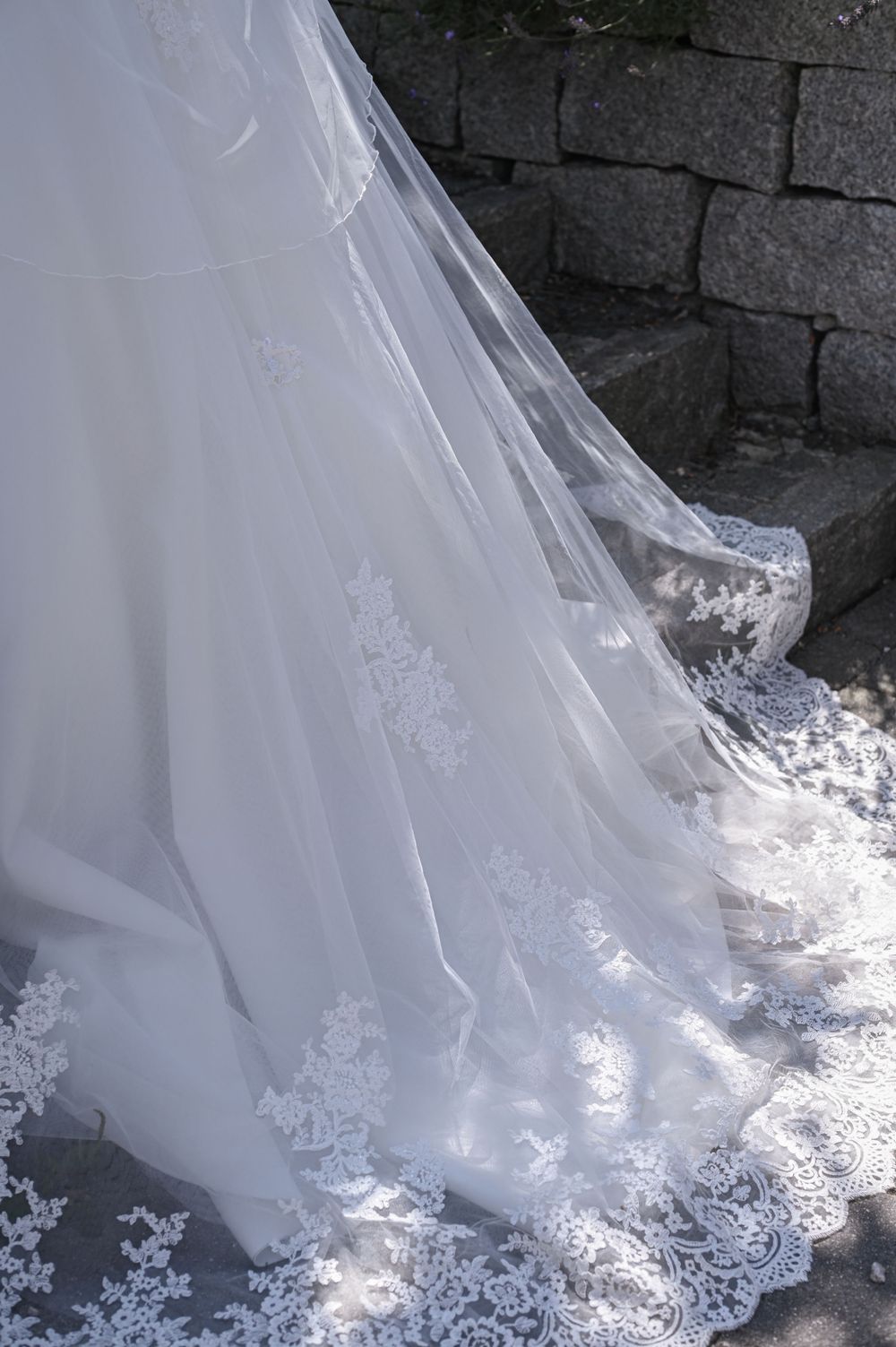 Zarte weiße Spitze säumt den Rand einer langen, fließenden Schleppe des Hochzeitskleides vor dem Hintergrund einer Steinmauer.