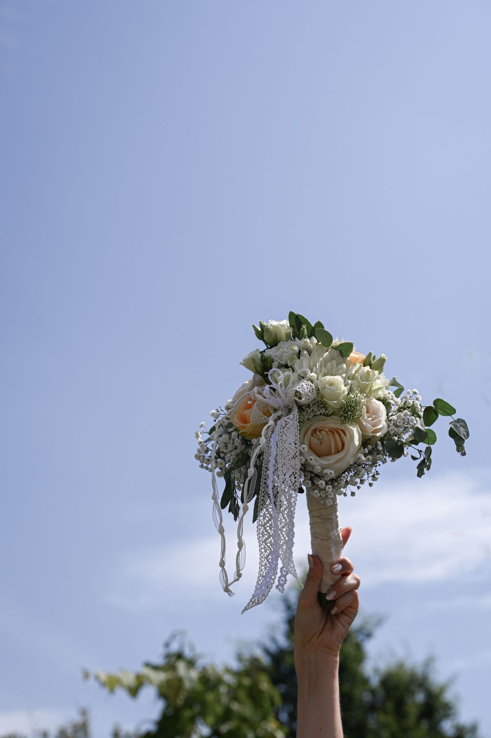 Eine Hand hält einen weißen Brautstrauß mit Rosen und Spitzenbändern vor dem Hintergrund eines blauen Himmels hoch.