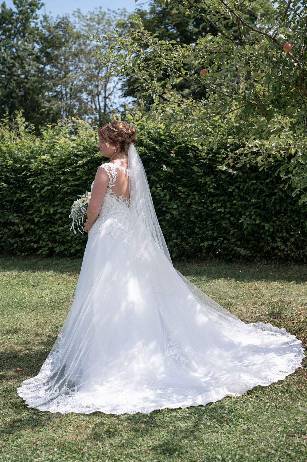 Ein Hochzeitskleid mit Spitzendetails und langer, fließender Schleppe wird von hinten in einer Gartenlandschaft gezeigt.