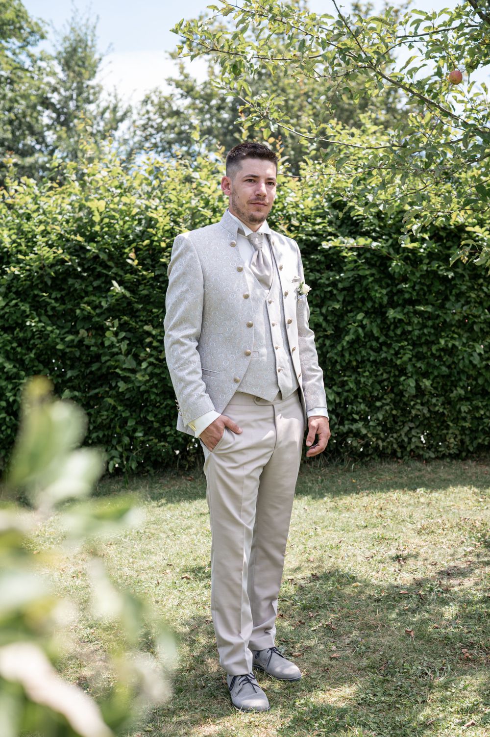 Elegantes cremefarbenes Anzug-Ensemble, fotografiert in einem Garten mit grünem Laub im Hintergrund.