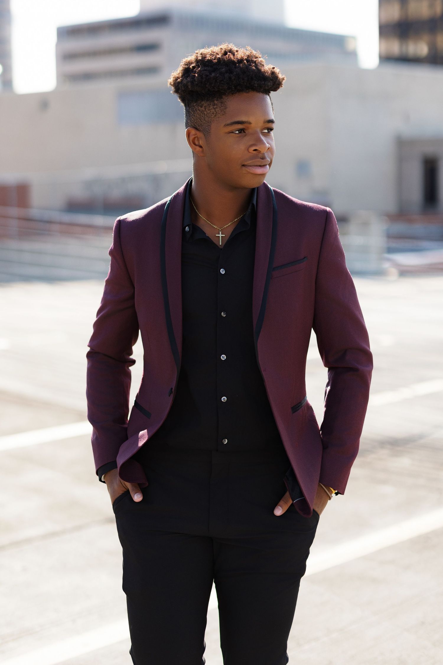 Young man wearing burgundy suit jacket and black shirt poses on urban rooftop.