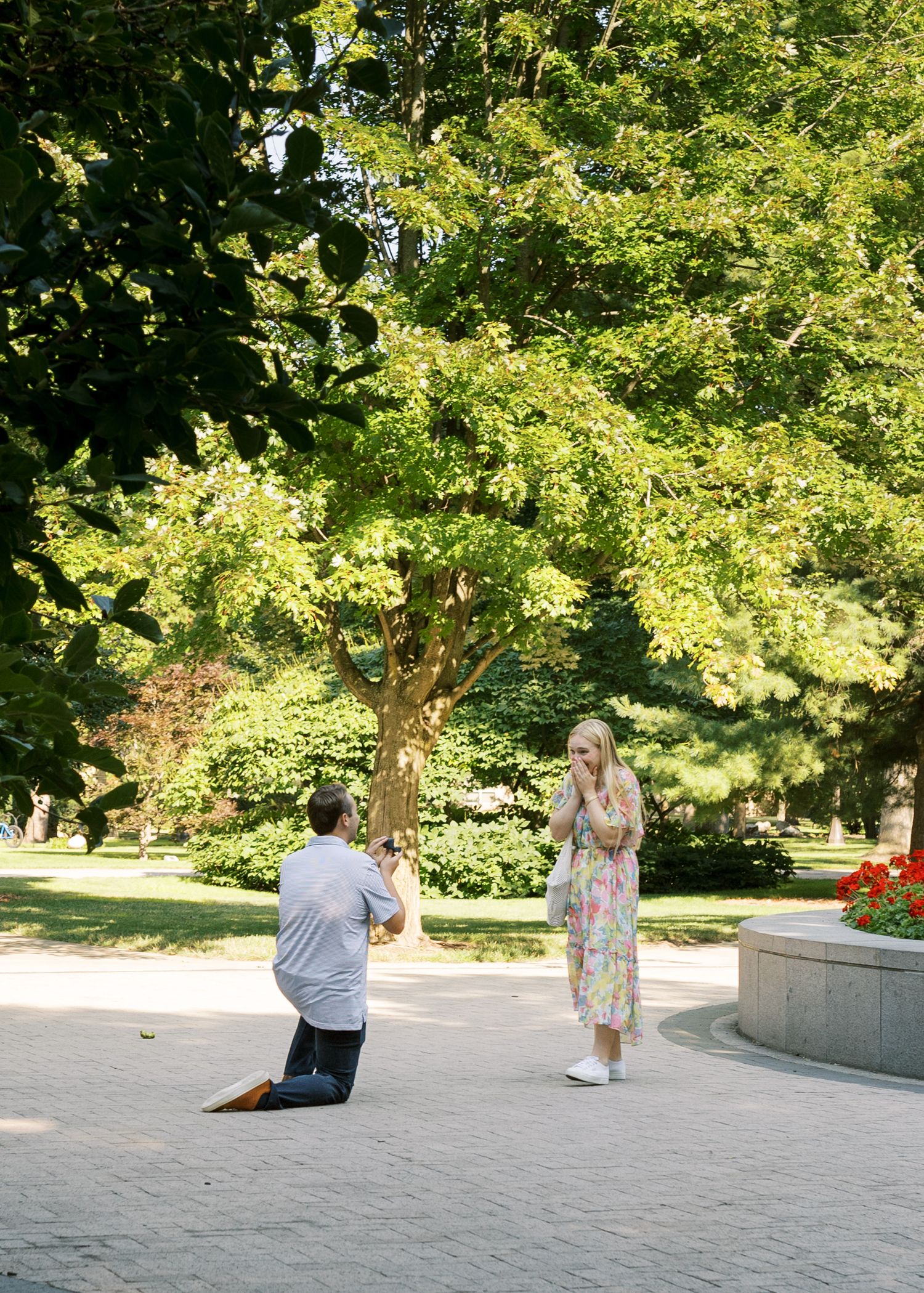 A surprise proposal moment captured on campus with lush green trees in the background.