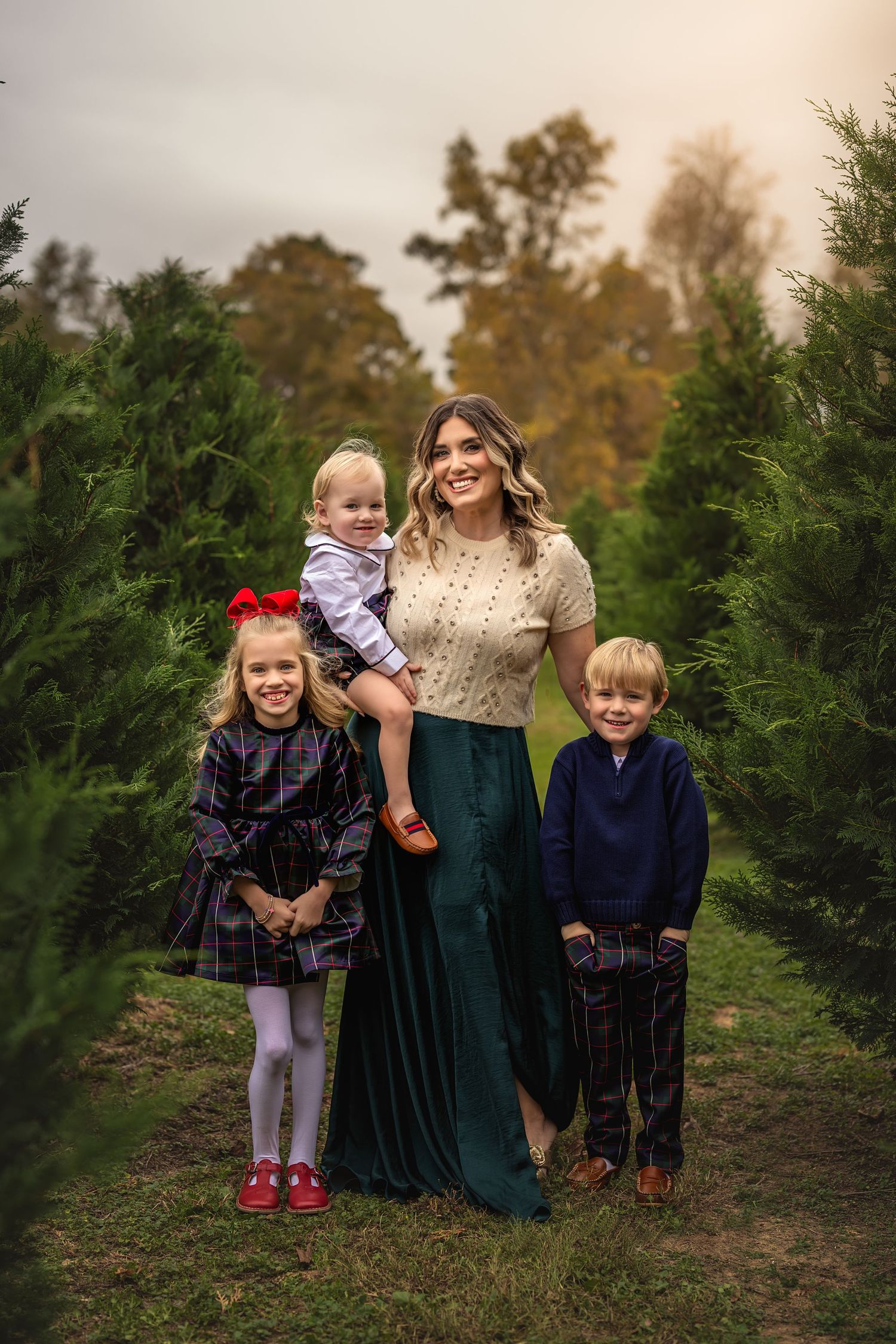 A mother poses with her children among evergreen trees at a Christmas tree farm.